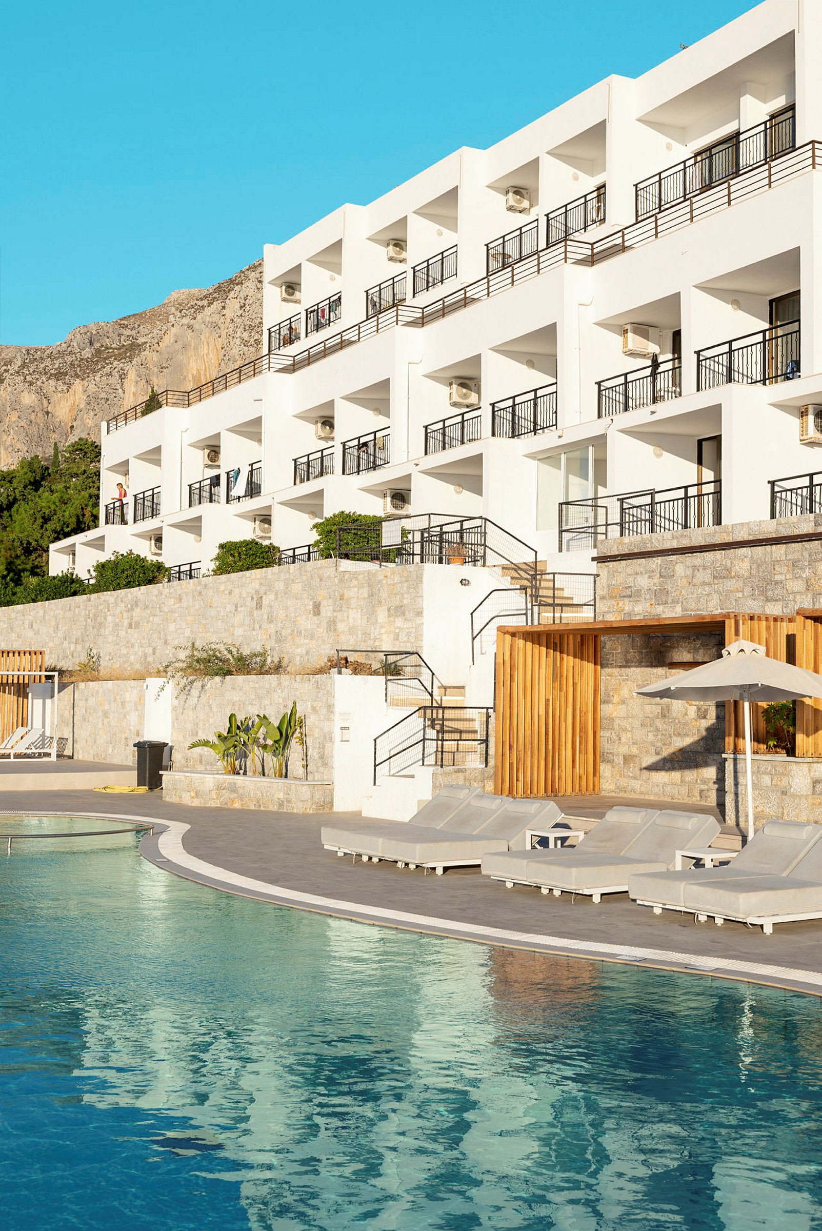 Ett hotell vid en pool med solstolar framför. Byggnaden har flera balkonger och syns omgiven av berg och klarblå himmel. (AI-genererad text)