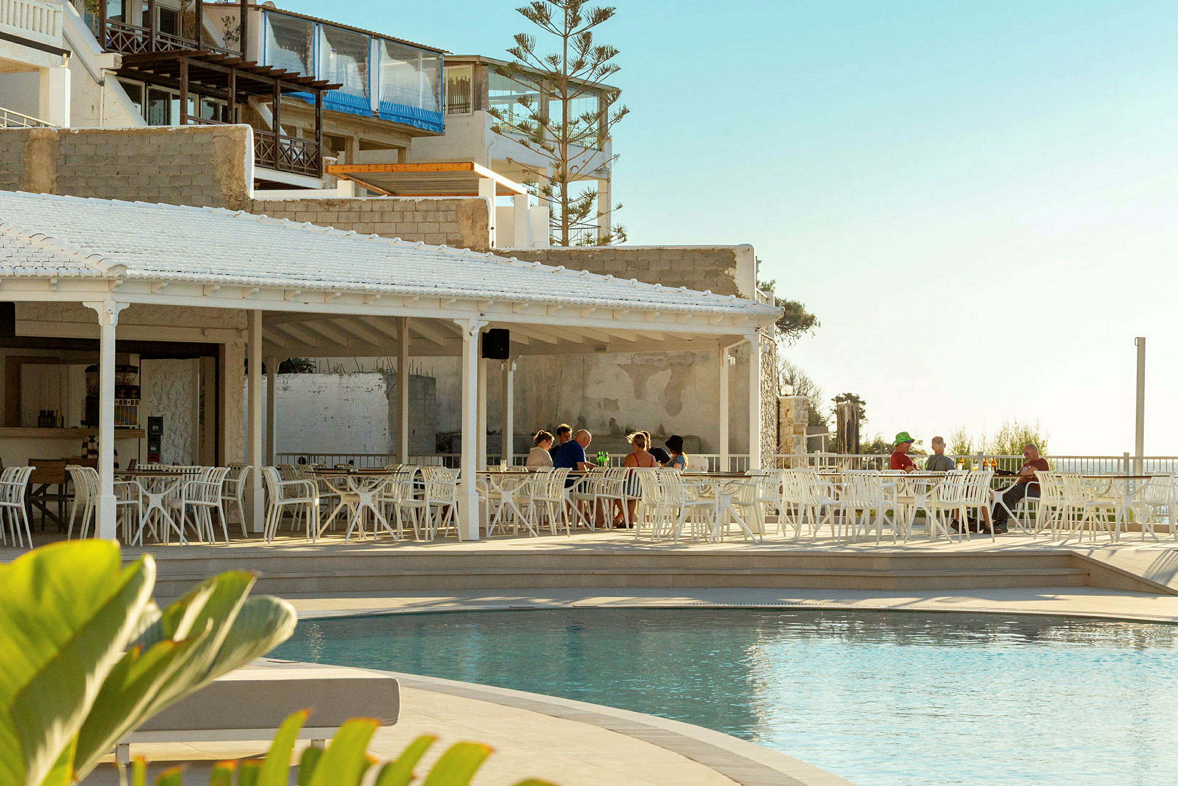 En utomhusrestaurang med vita möbler vid sidan av en pool, med några personer som sitter vid borden. Bakgrunden består av byggnader och en ljus himmel. (AI-genererad text)