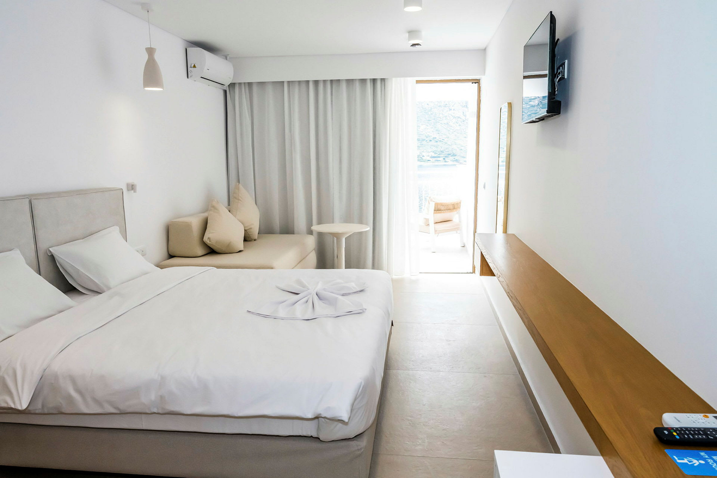 Ett hotellrum med en säng, soffa, ett bord och en TV på väggen. Ett fönster med ljusa gardiner leder ut till en terrass. (AI-genererad text)