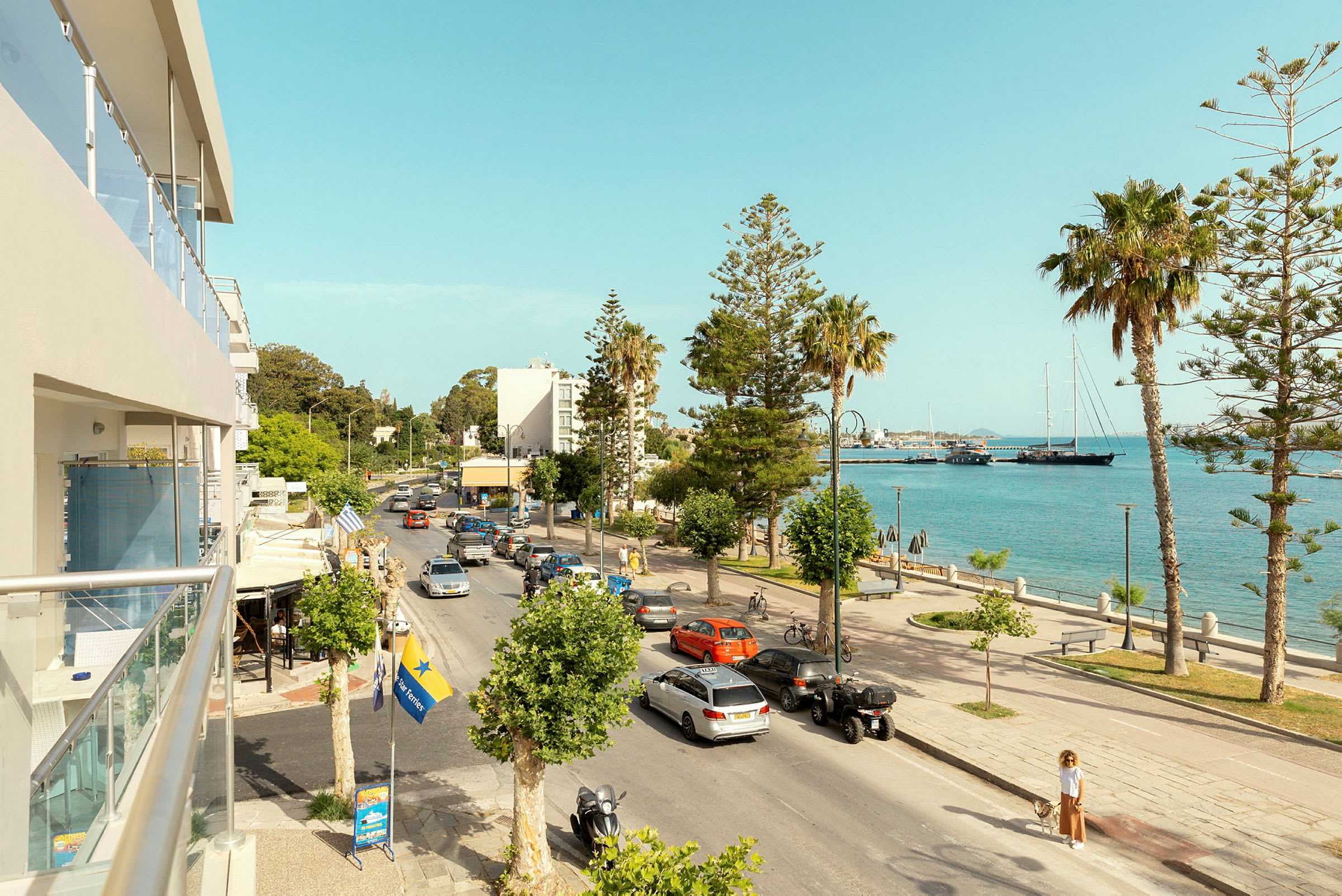 En stadsgata som går bredvid havet, med bilar, palmer och några byggnader i bakgrunden. (AI-genererad text)