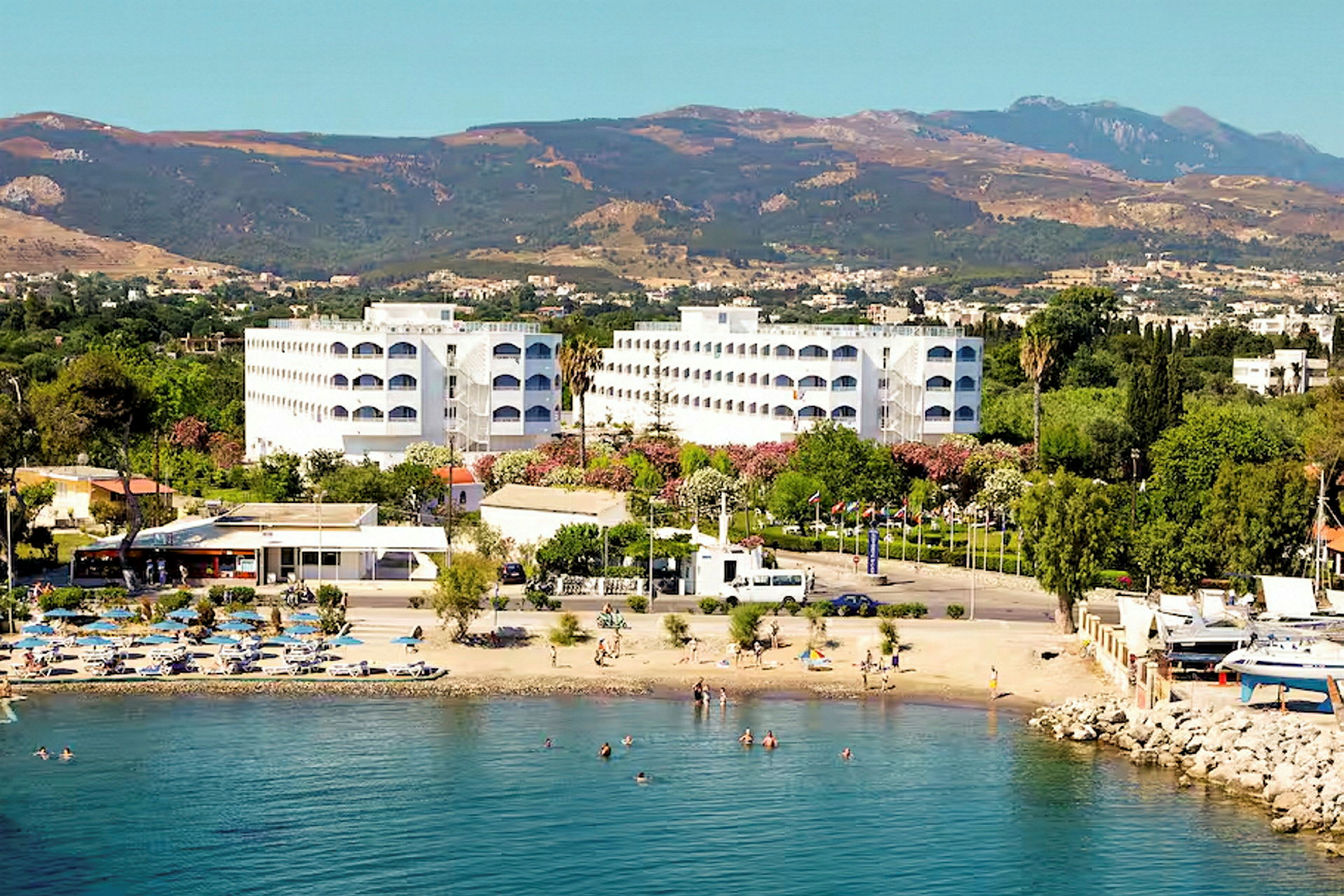 Hotellbyggnader vid en strand med badande personer och berg i bakgrunden. (AI-genererad text)