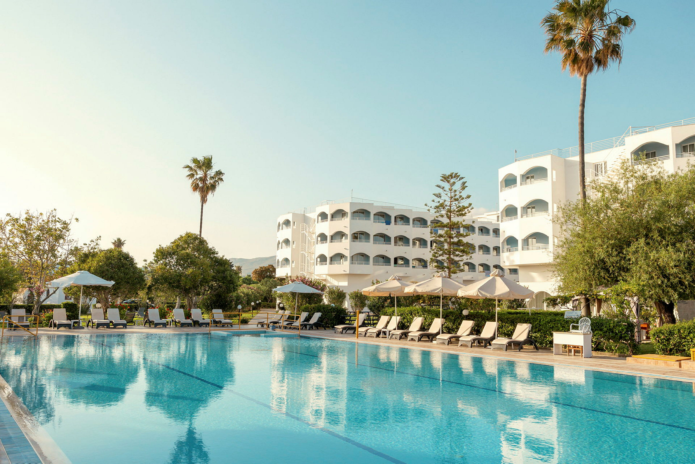 Hotellbyggnad med pool, solstolar och parasoller i förgrunden. (AI-genererad text)