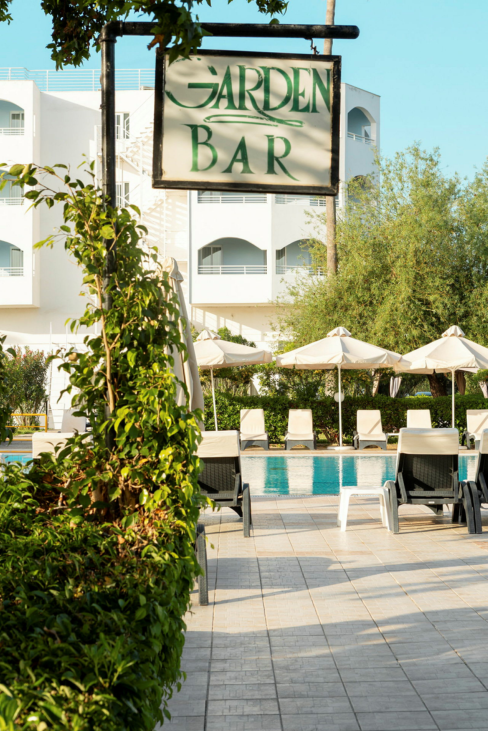 Skylt som visar 'Garden Bar' framför poolområde och hotellbyggnad. (AI-genererad text)