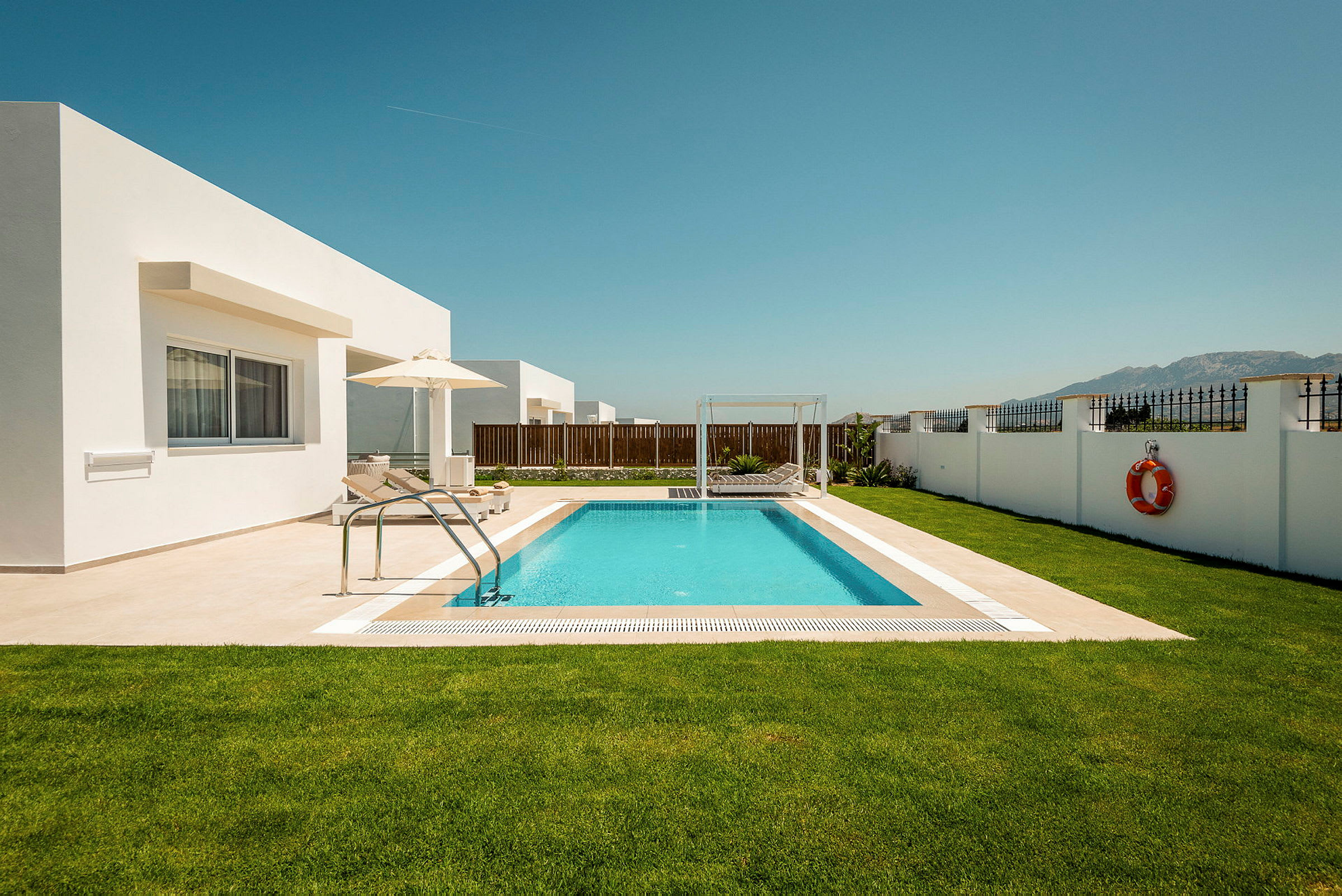 En privat pool med solstolar och parasoll omgiven av en gräsmatta och en vit mur. Modern arkitektur och klarblå himmel. (AI-genererad text)