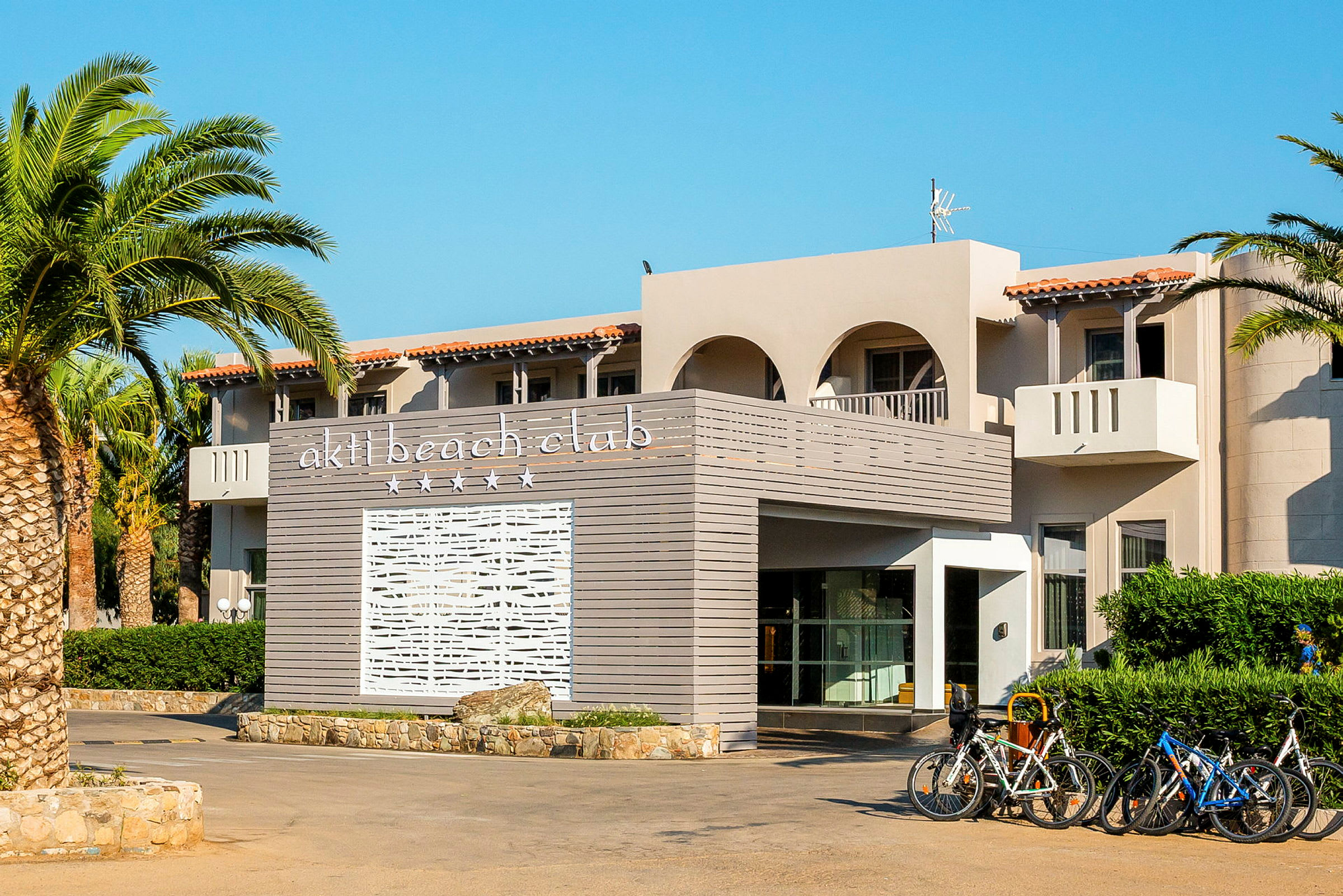 Byggnad med texten 'akti beach club' på en panel i förgrunden och cyklar parkerade vid entrén. (AI-genererad text)