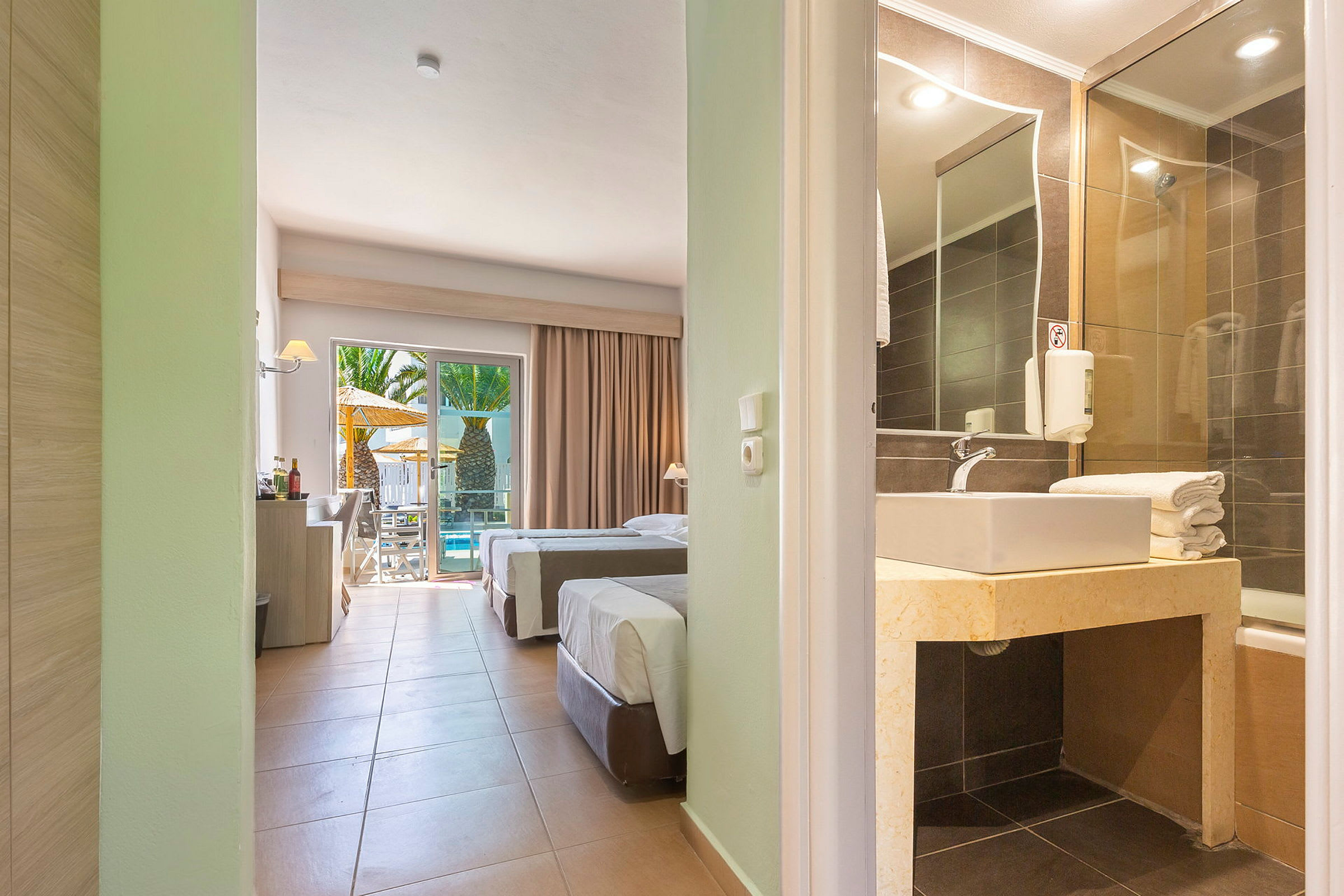Bild av ett hotellrum med två enkelsängar, ett badrum med handfat och en uteplats med pool och palmer. (AI-genererad text)