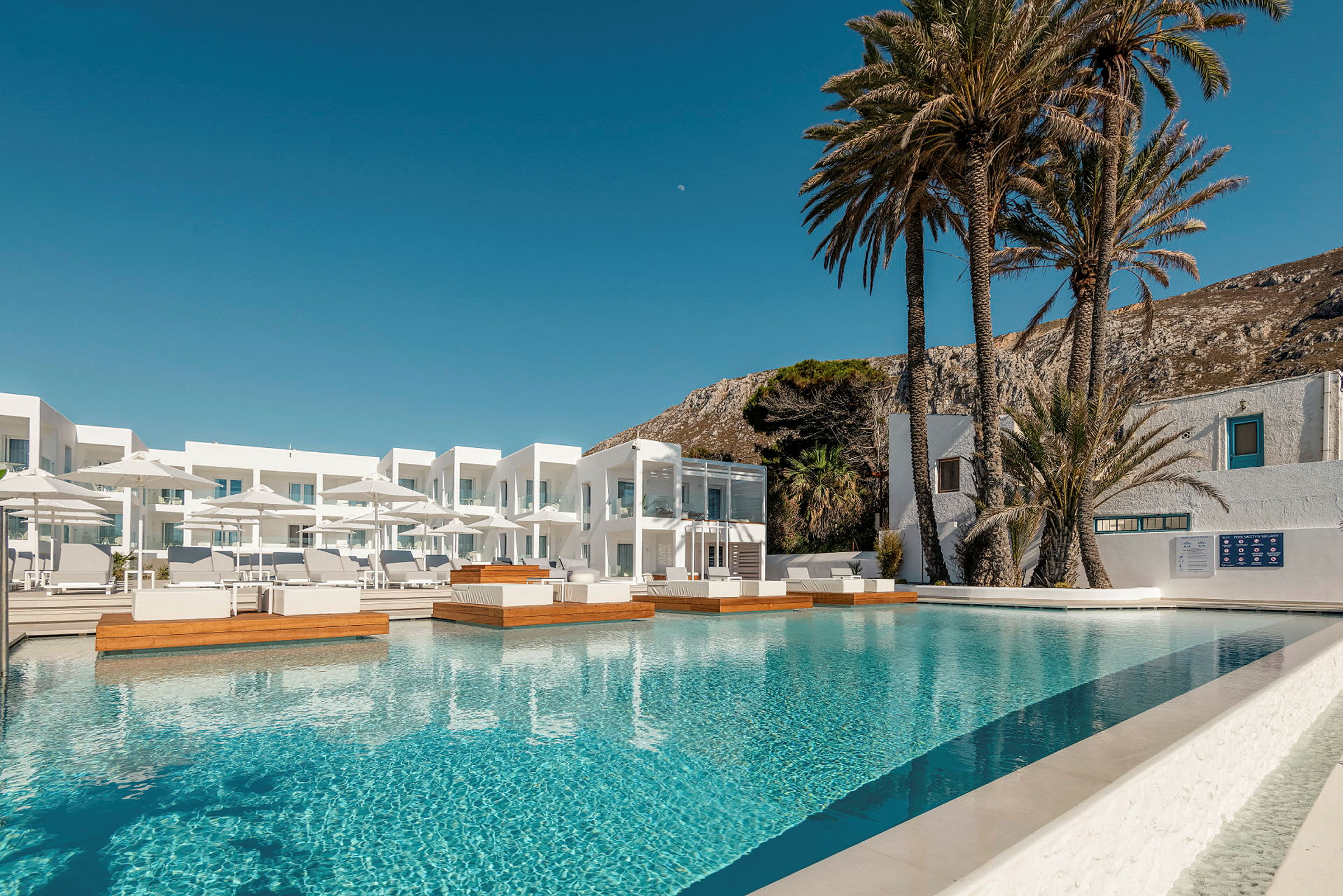 Ett hotellområde med en pool, solstolar och parasoll, omgärdat av vita byggnader och palmer. (AI-genererad text)