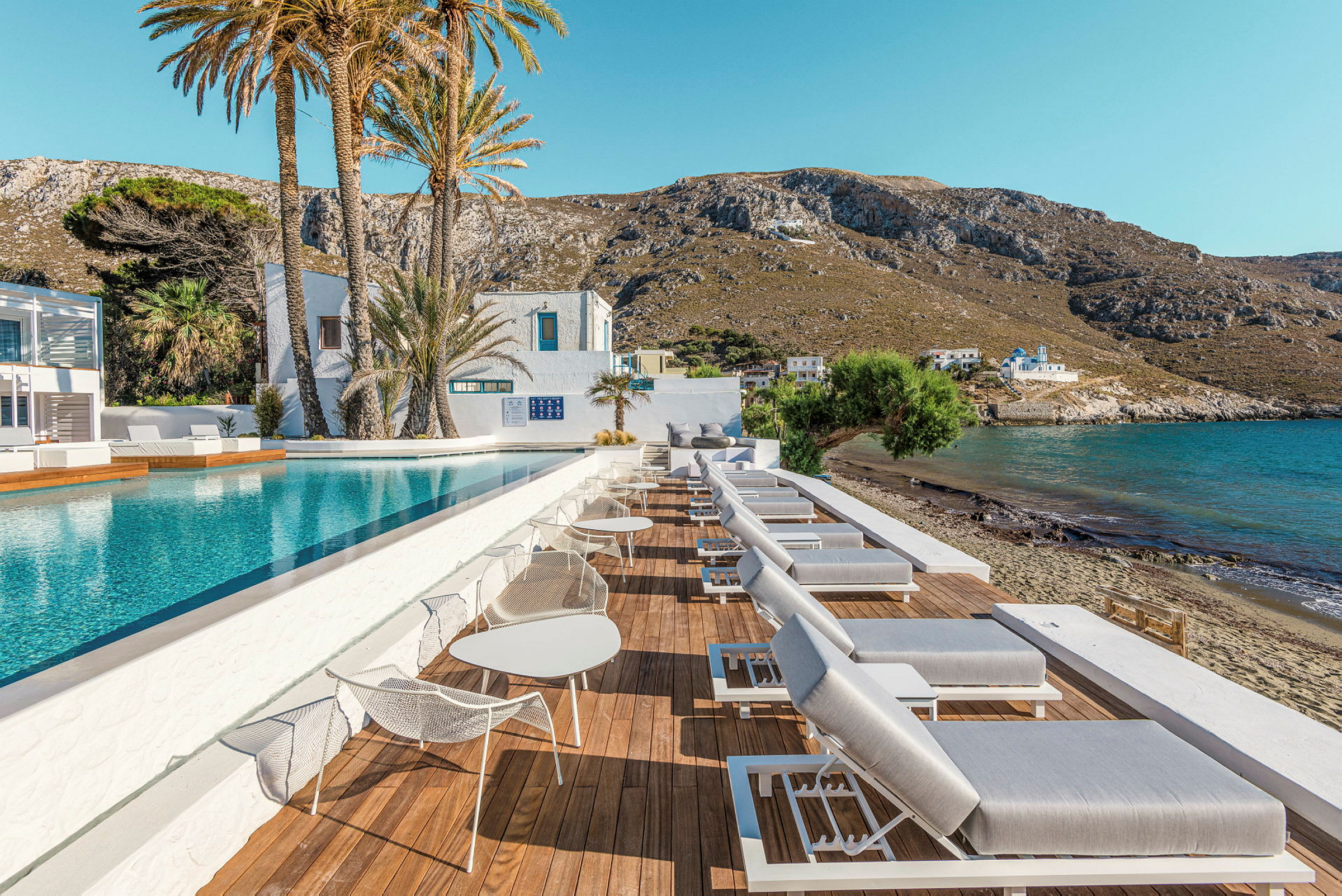 Utomhuspool med relaxstolar och utsikt över havet och berg i bakgrunden. (AI-genererad text)