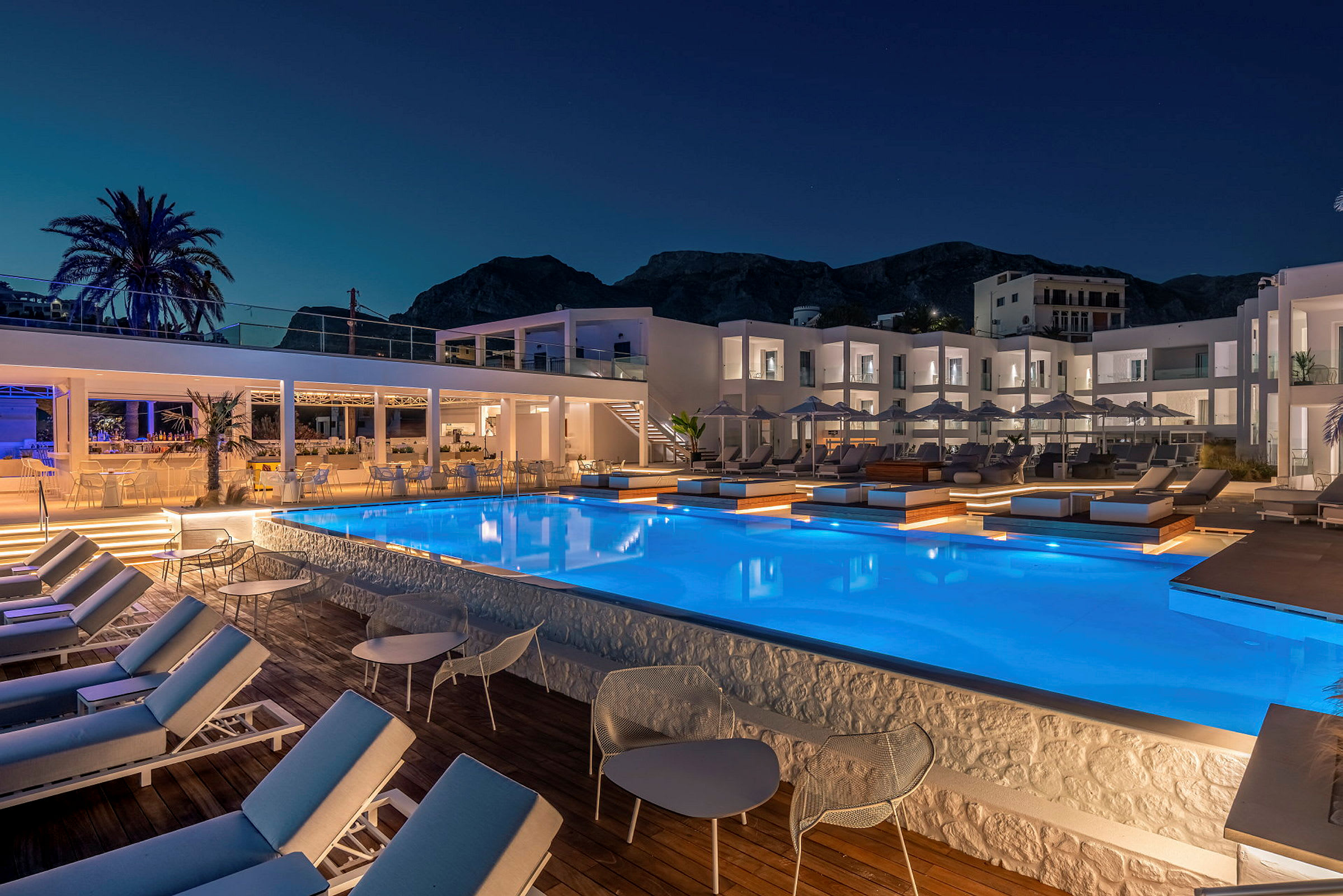 Bild på en upplyst hotellpool och omgivande solsängar och byggnader i kvällsmörker. (AI-genererad text)