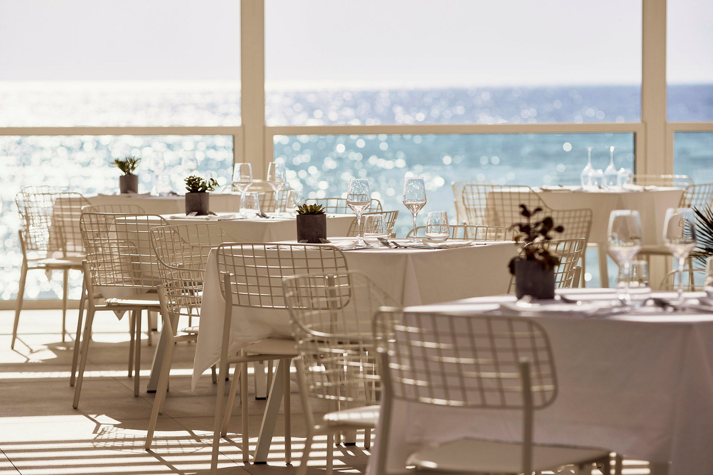 Restaurangbord med vita dukar och stolar nära ett stort fönster med utsikt över havet. (AI-genererad text)