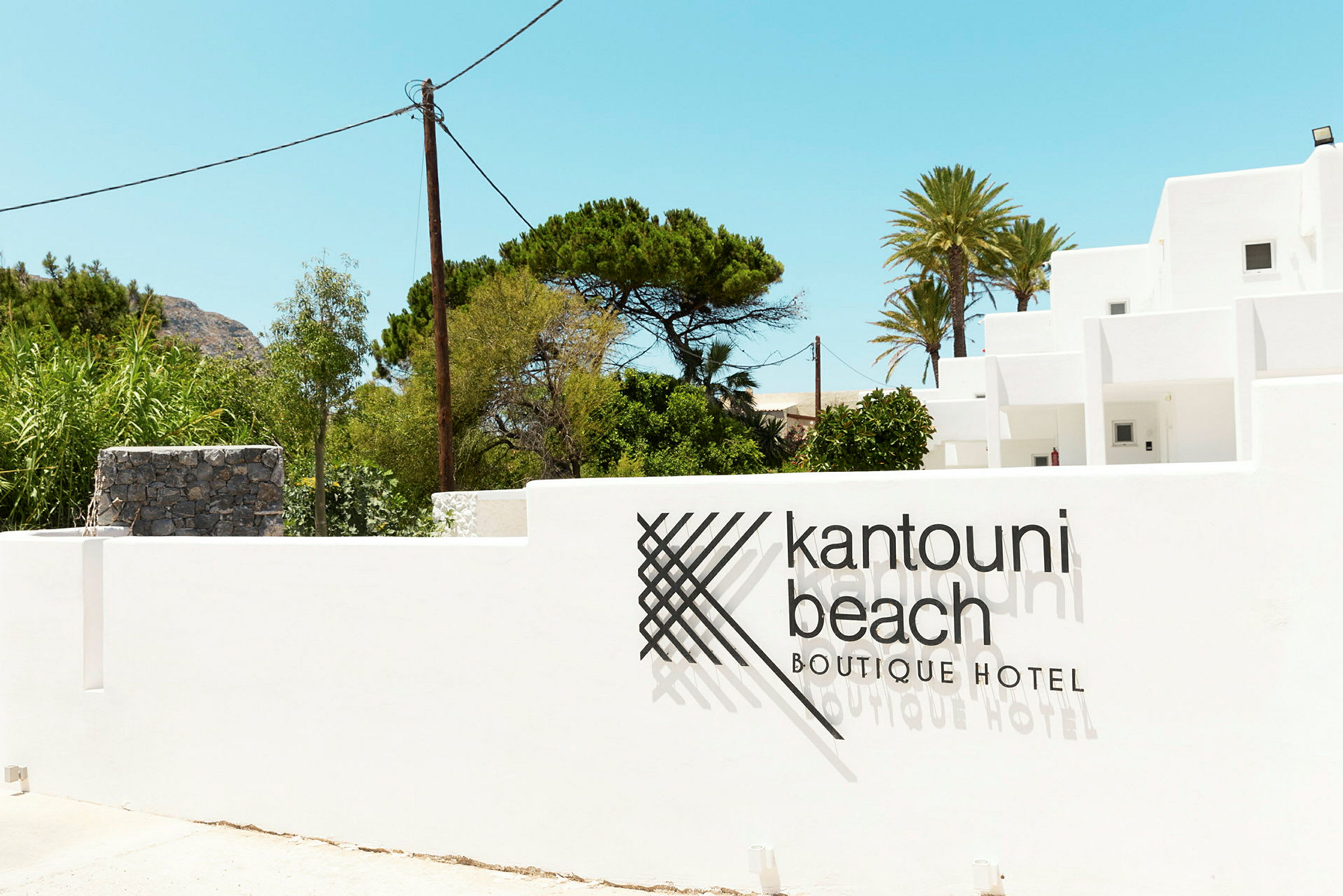 Bild på en vit byggnad med texten 'Kantouni Beach Boutique Hotel' på muren framför. Grönska och klar himmel i bakgrunden. (AI-genererad text)