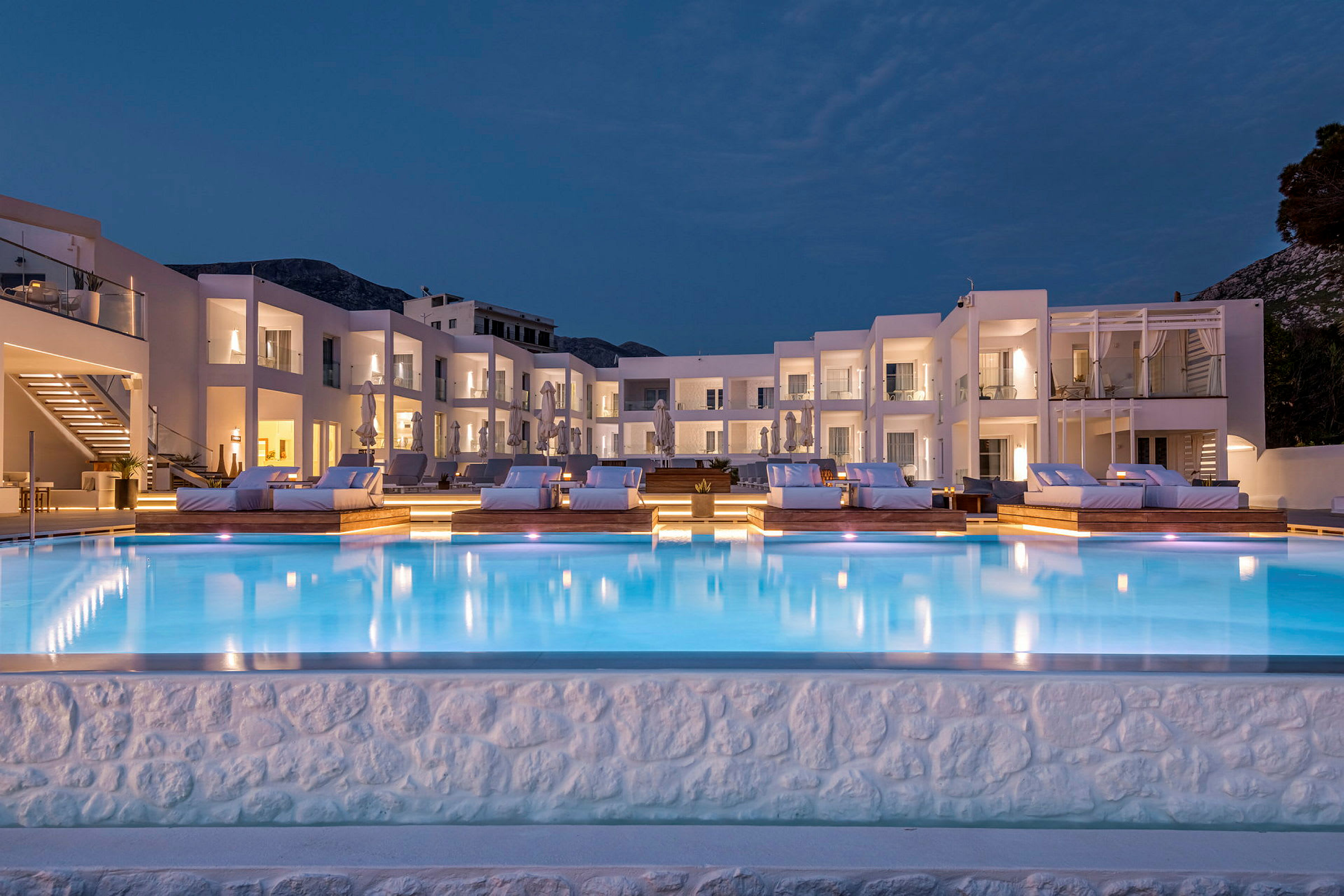 Upplyst hotellpool med solstolar och vita byggnader i bakgrunden. (AI-genererad text)