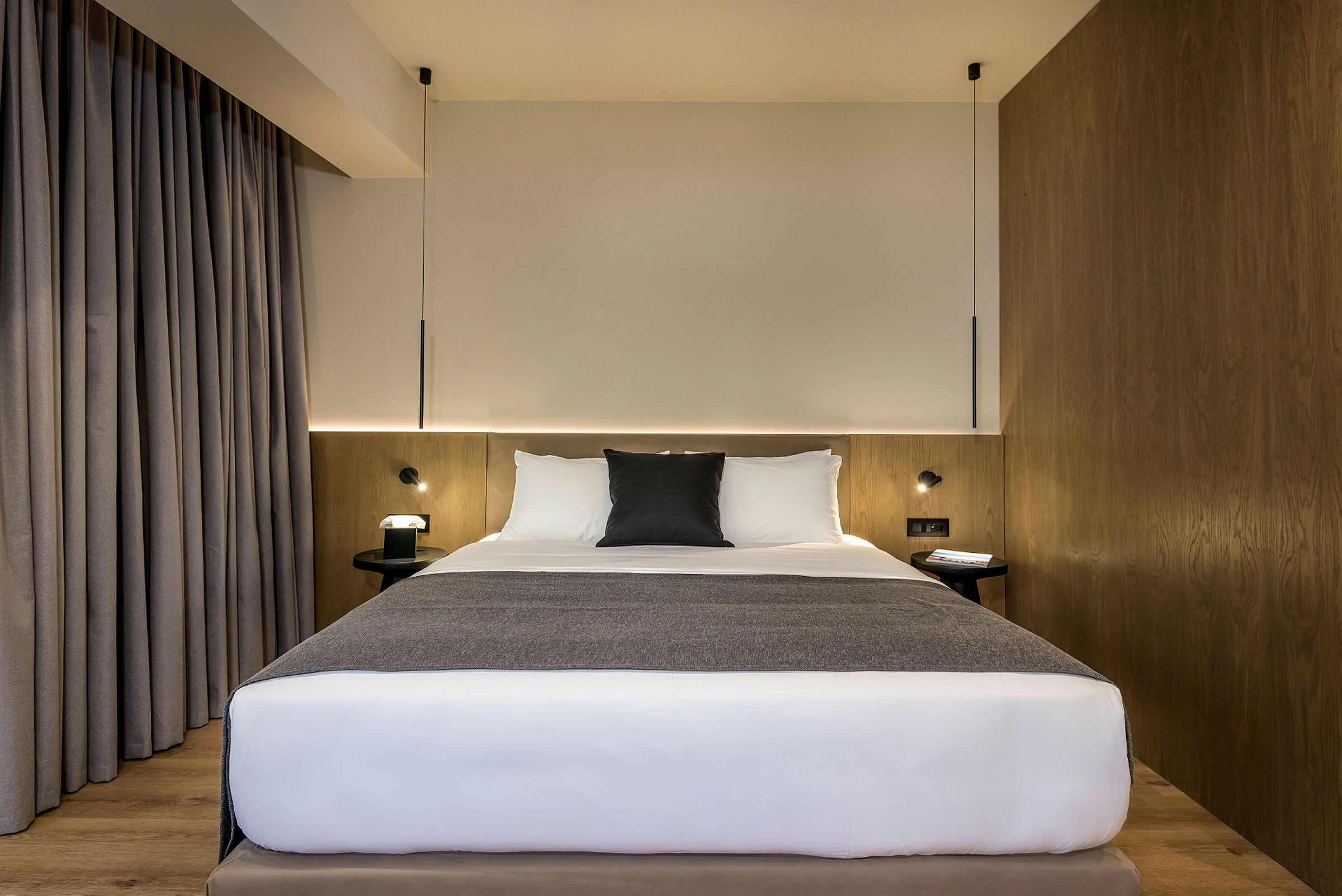 Ett hotellrum med en säng med vita lakan och gråa detaljer, träpanel och gråa gardiner. (AI-genererad text)
