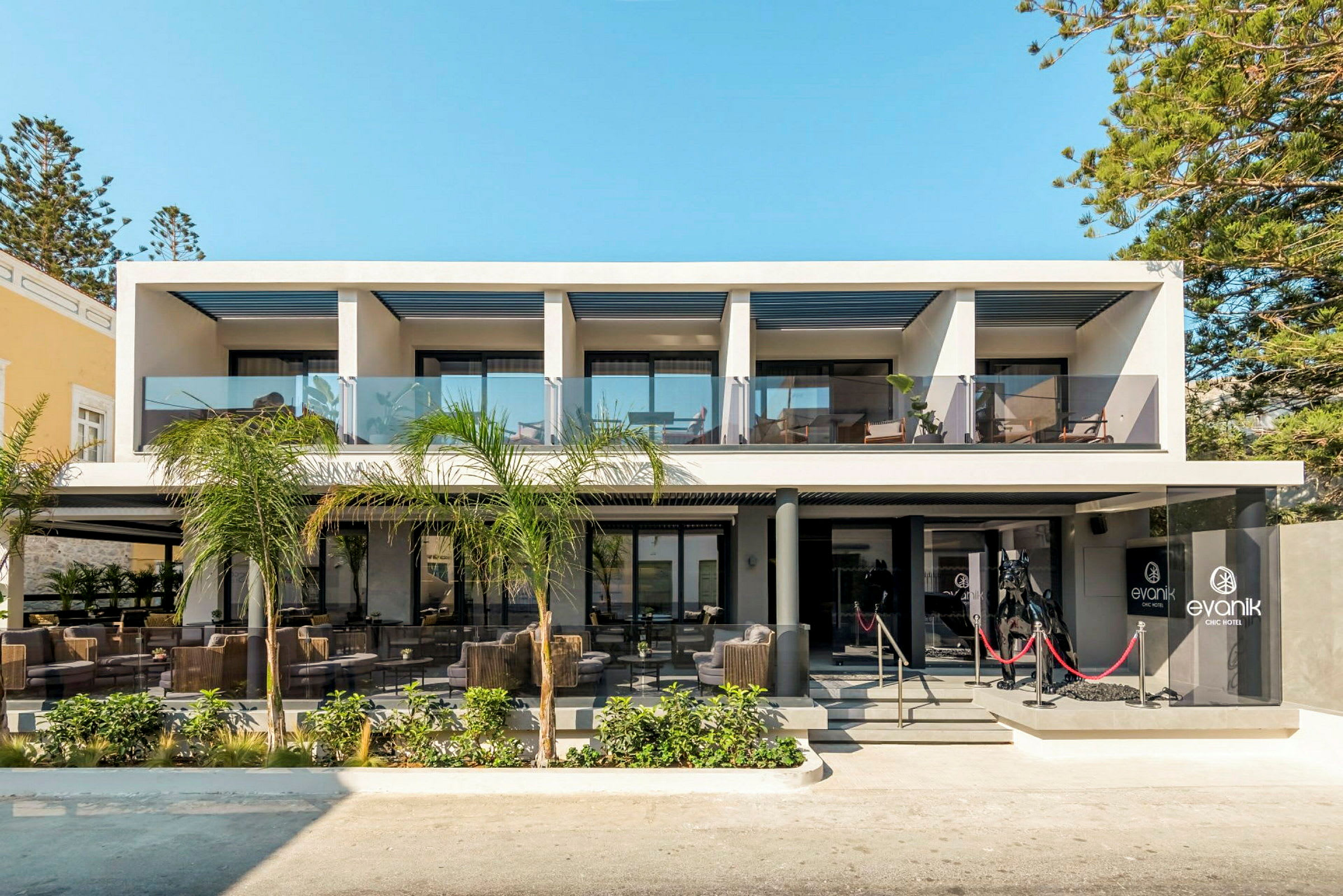 Modern hotellbyggnad med glasräck och utemöbler vid entrén. (AI-genererad text)