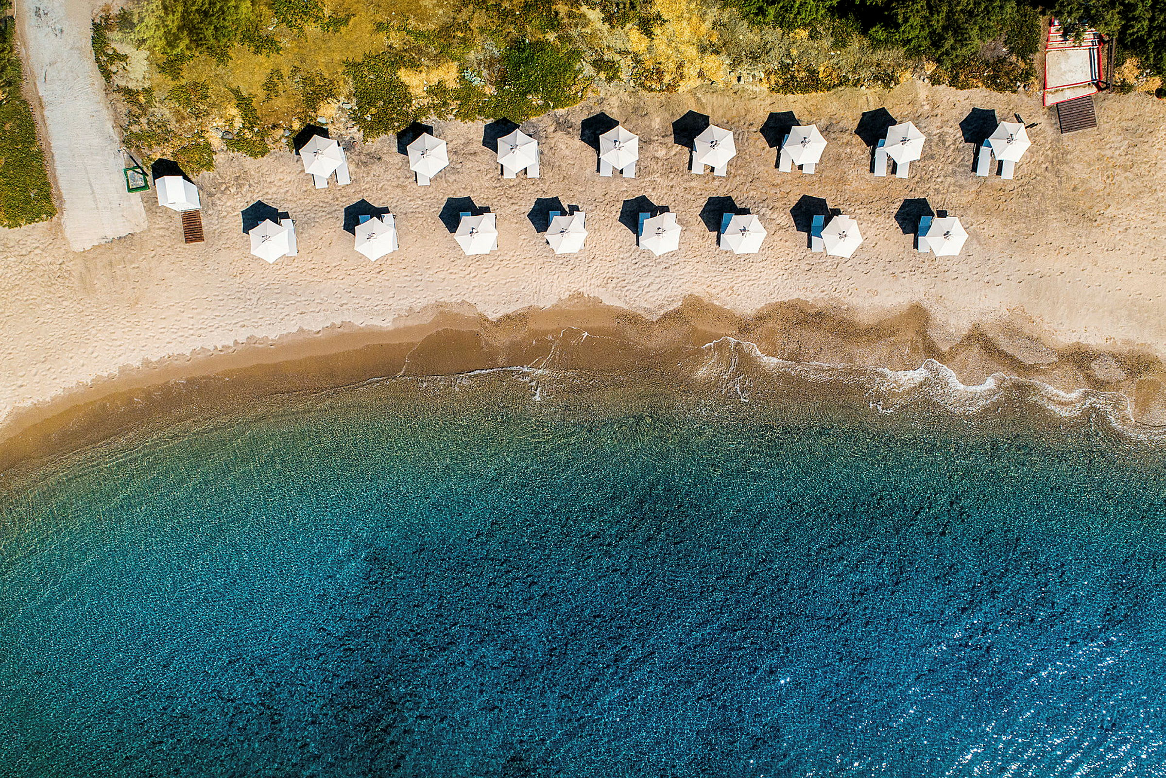 Flygfoto av en sandstrand med parasoller i raka rader och klart blått vatten. (AI-genererad text)