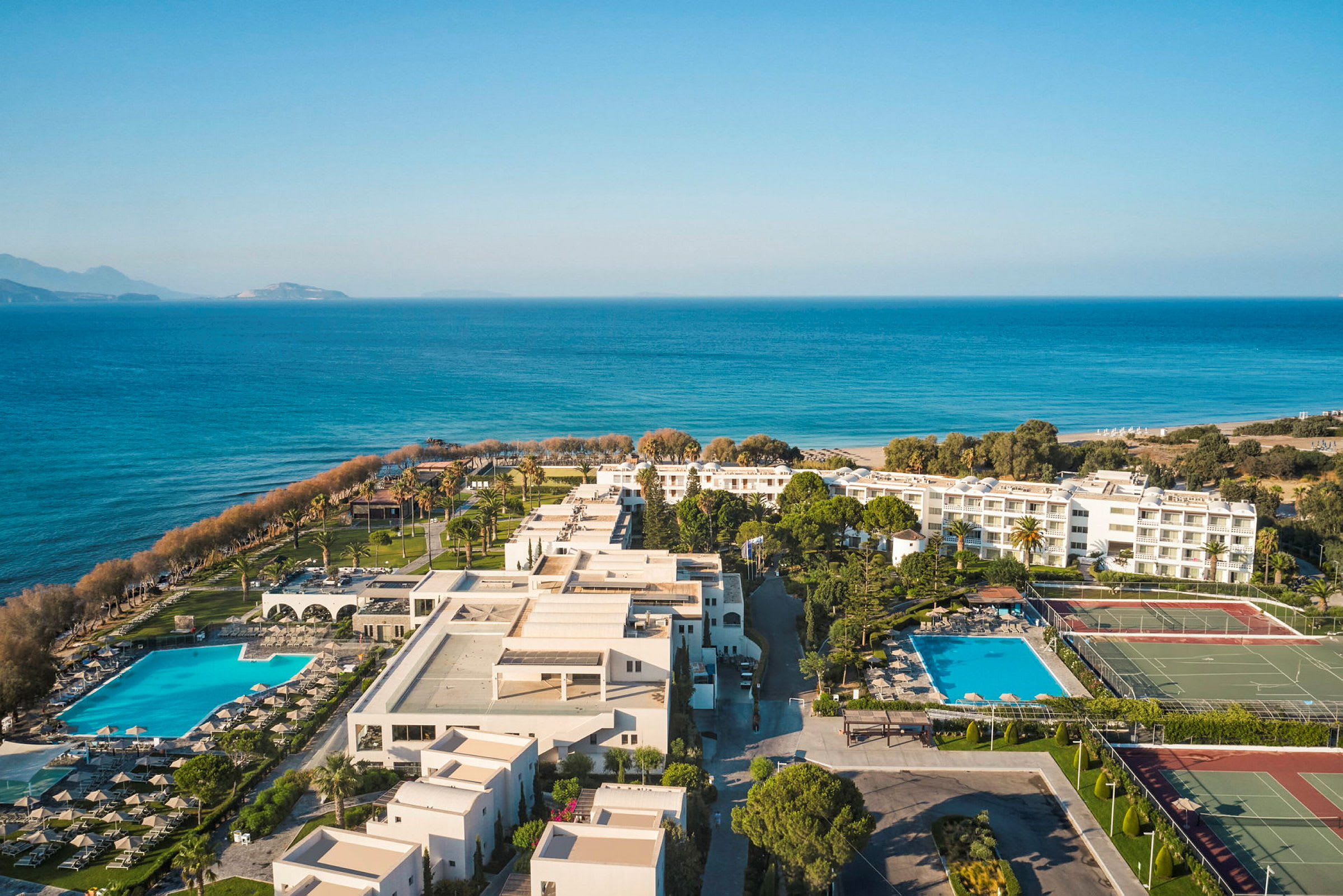 Flygfoto av en semesteranläggning vid havet med swimmingpooler, byggnader och tennisbanor. (AI-genererad text)
