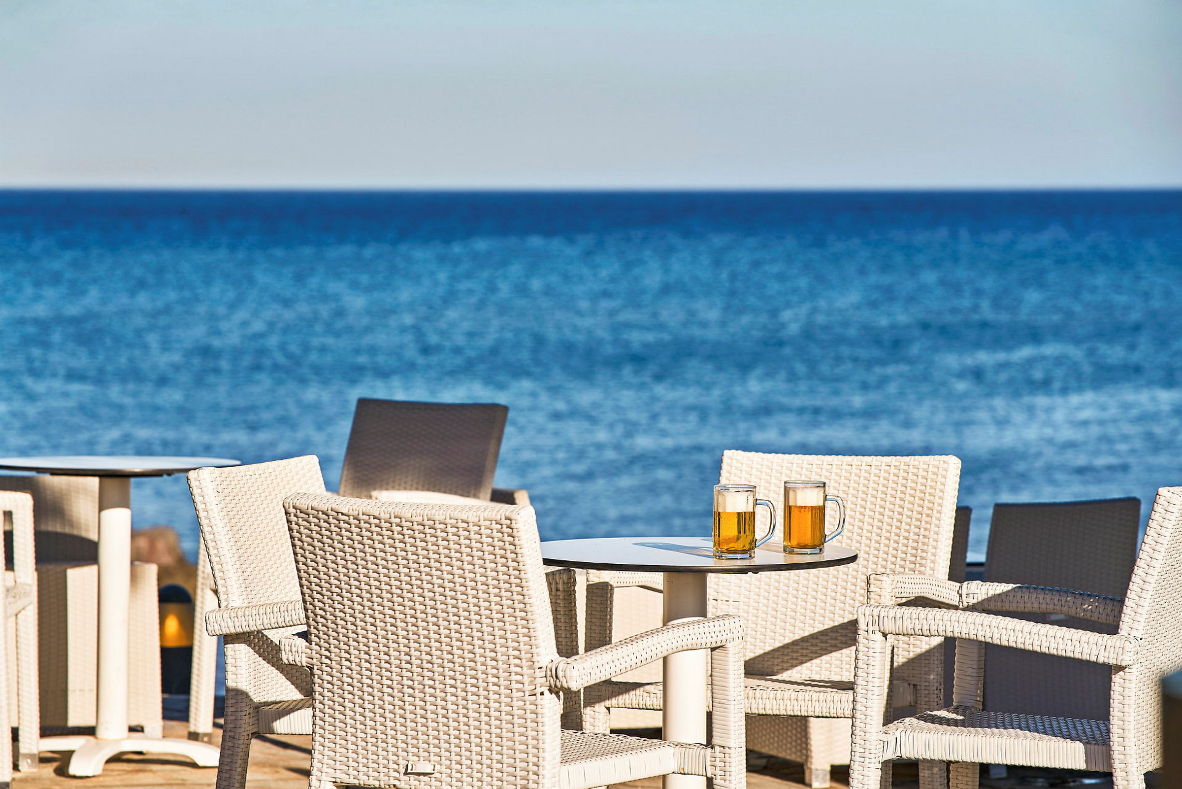 Korgstolar vid ett bord med två ölglas, med havet i bakgrunden. (AI-genererad text)