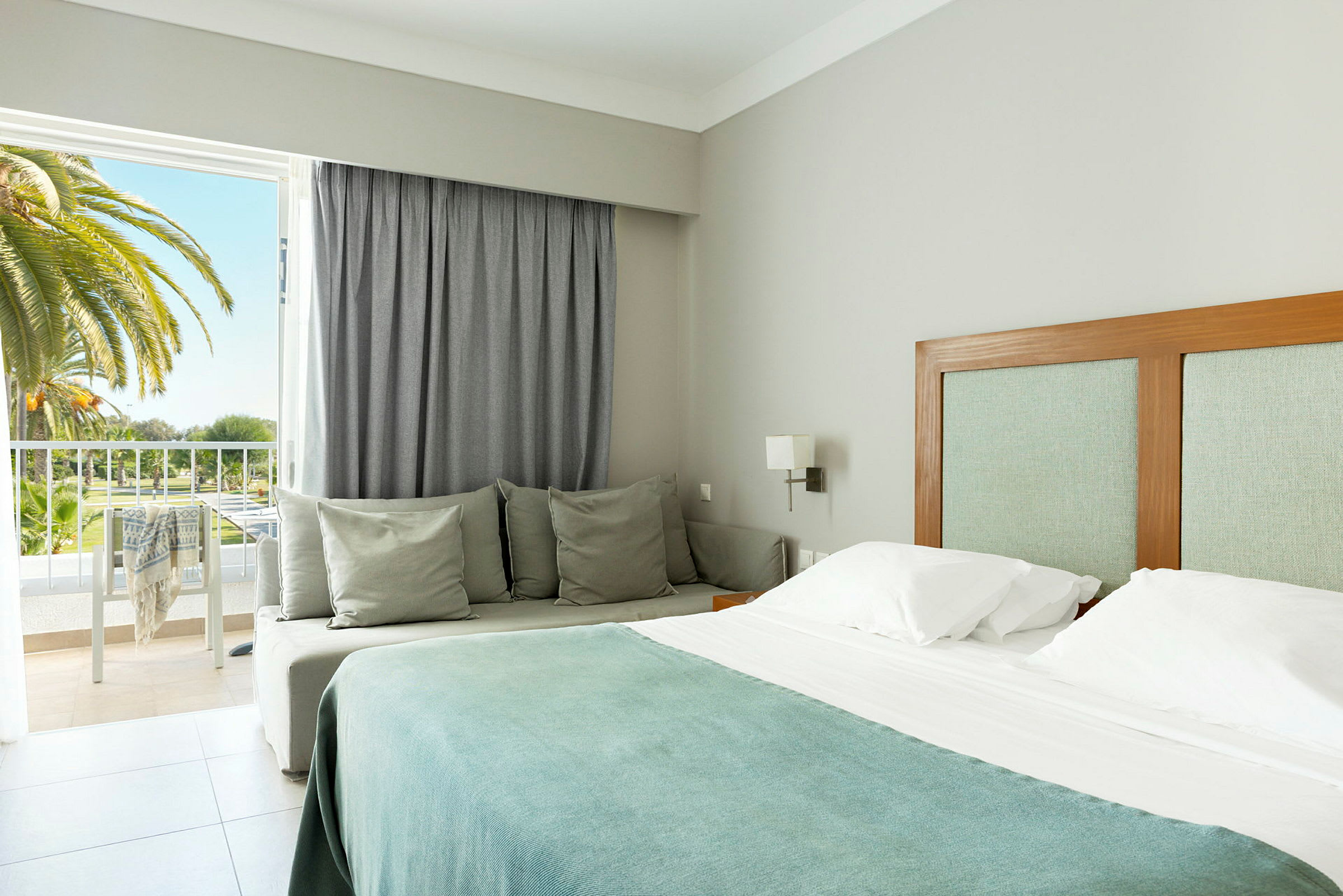 Ett hotellrum med en dubbelsäng, en soffa och en balkong med utsikt över palmer. (AI-genererad text)