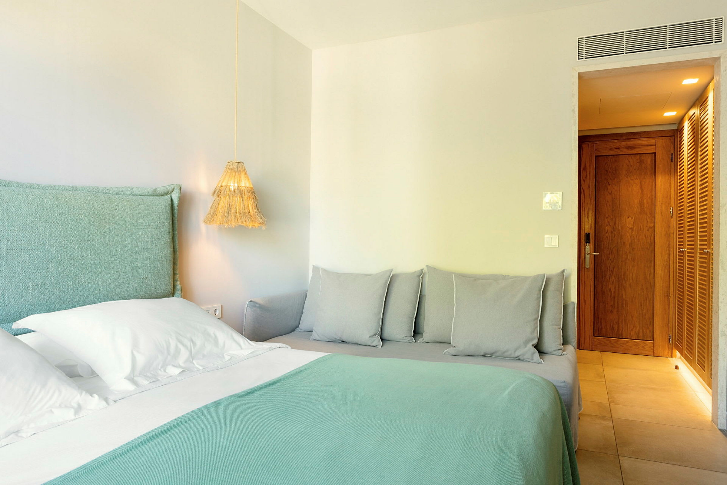 Ett hotellrum med en säng med turkosa lakan och vitkudde, samt en närliggande soffa med grå kuddar. (AI-genererad text)