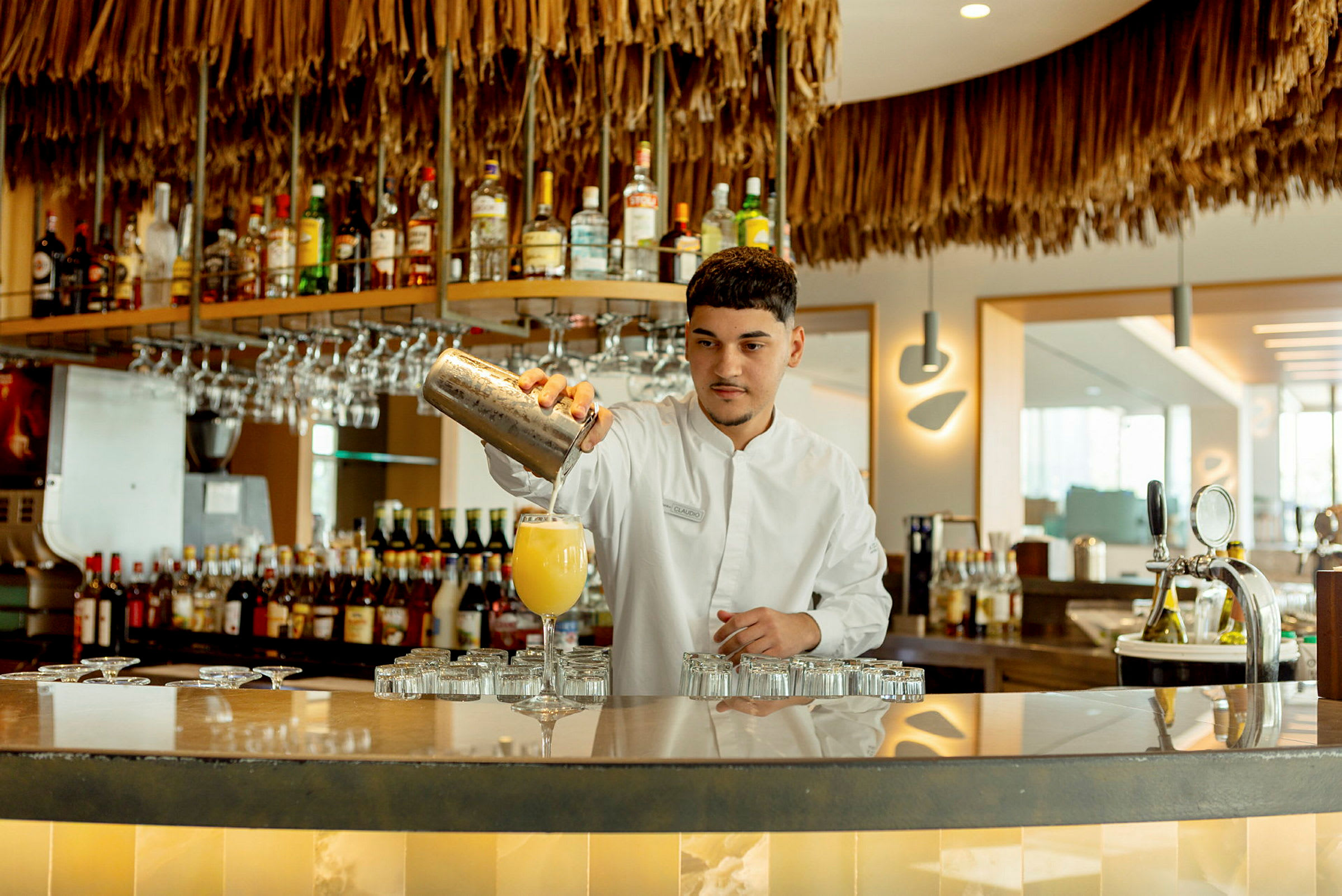 En bartender häller drycken i ett glas framför en hylla med olika spritflaskor och vinglas. (AI-genererad text)