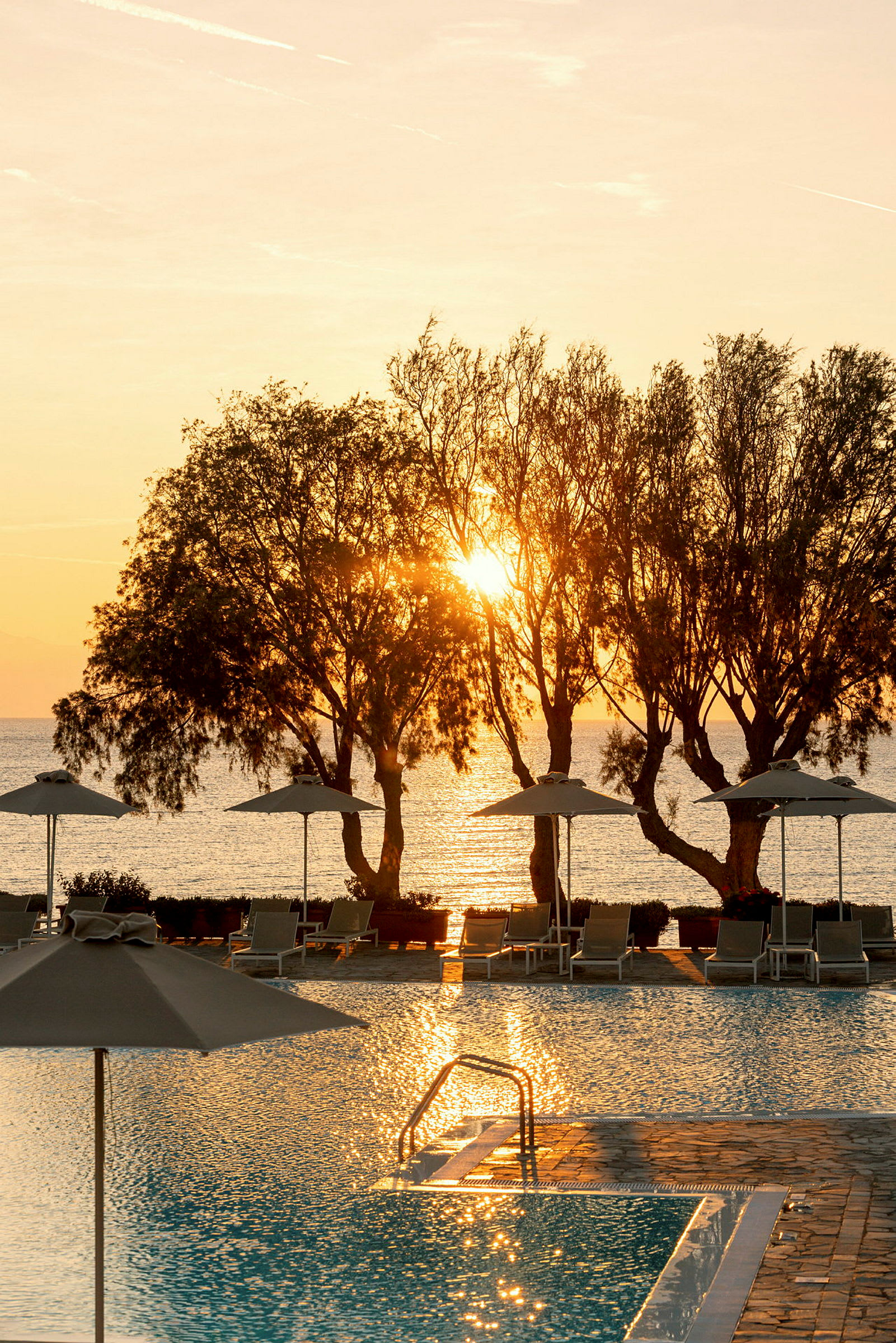 Bild av en pool med stolar och parasoll, framför träd och en solnedgång vid havet. (AI-genererad text)