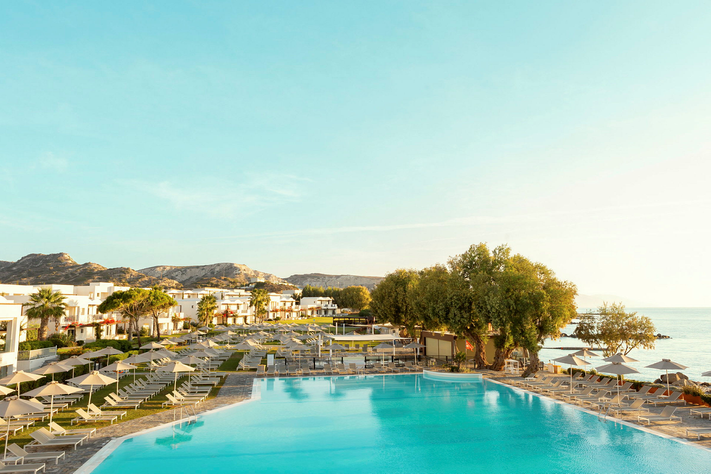 Vy över en stor pool vid ett hotell, omgiven av solstolar och parasoller, med utsikt över havet och omgivande landskap. (AI-genererad text)