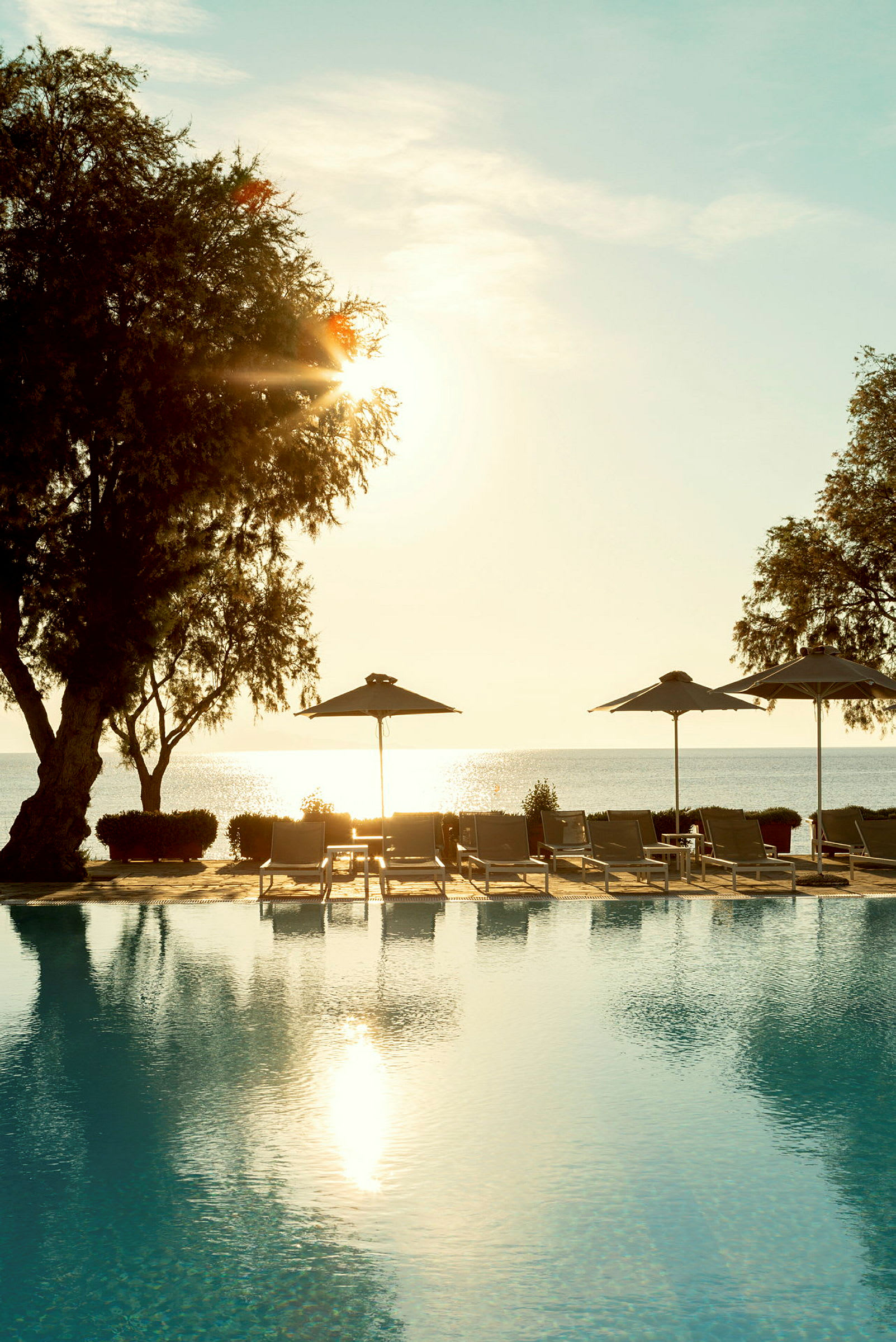 En solnedgång sedd över en pool bredvid stranden, med solstolar och parasoller i förgrunden. (AI-genererad text)