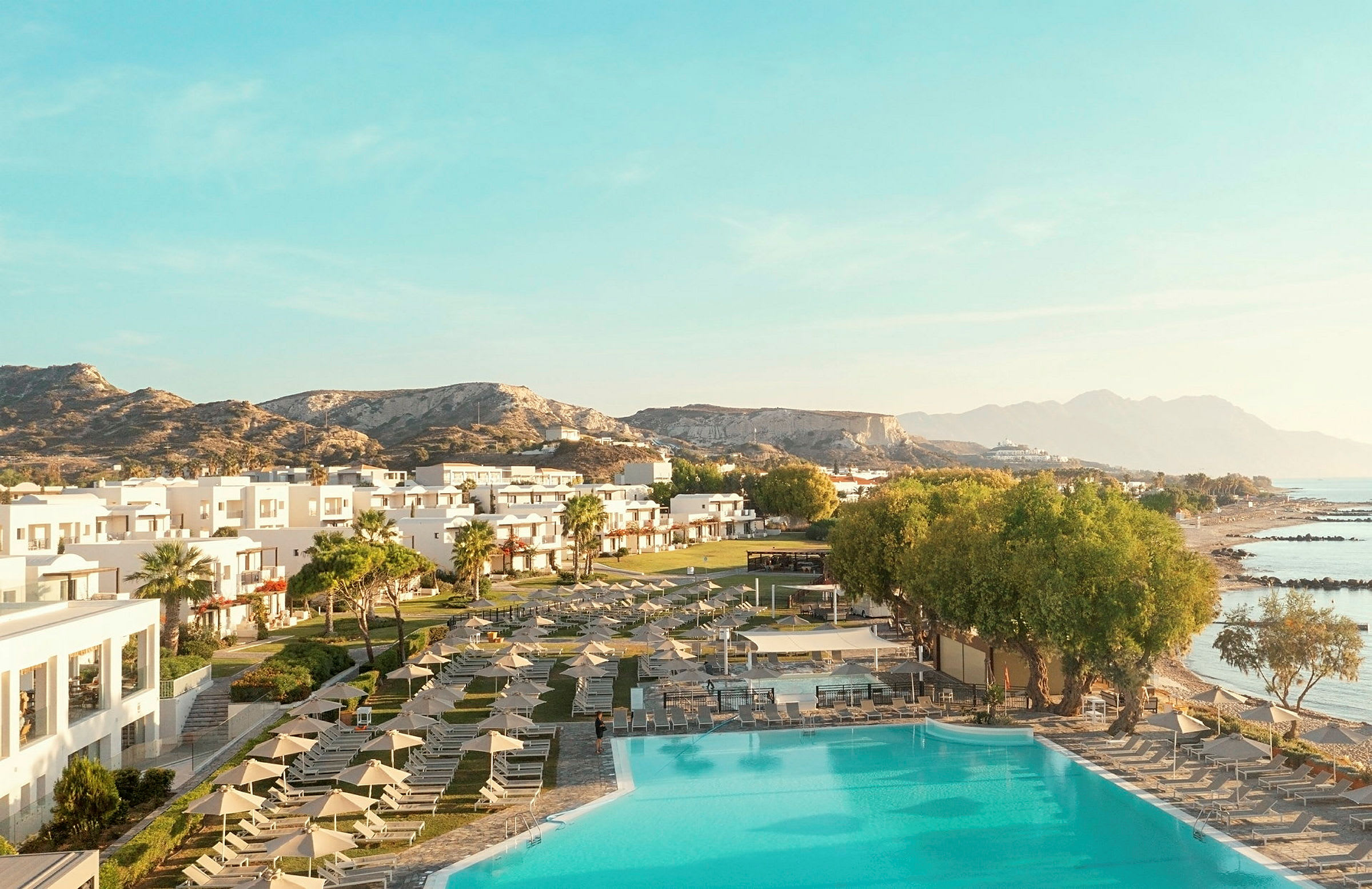 Vy över ett hotellområde med pool, grönområden och sandstränder, omgivet av berg och hav. (AI-genererad text)