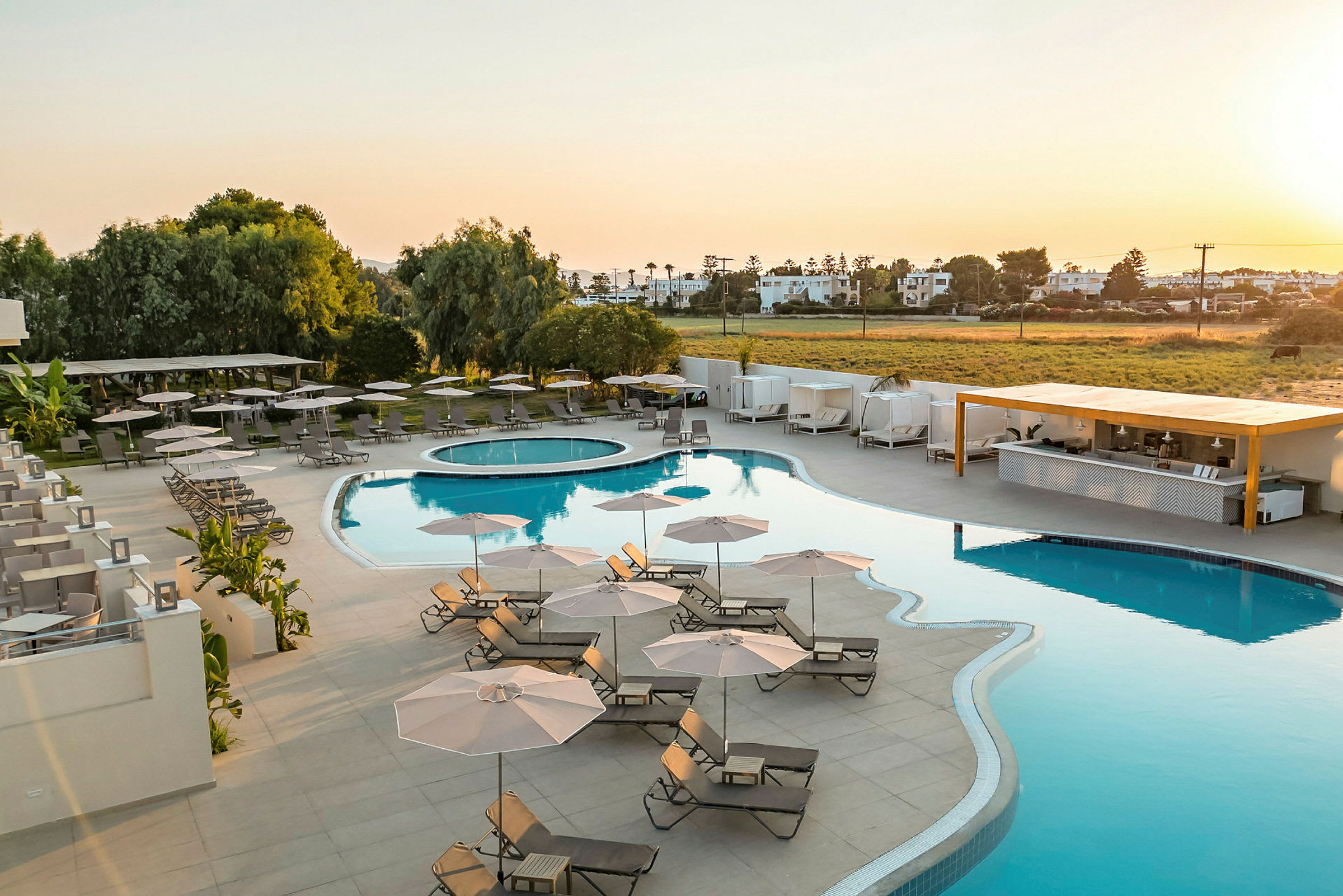 Ett hotellområde med en stor pool omgiven av solstolar och parasoller, med grönska och byggnader i bakgrunden. (AI-genererad text)