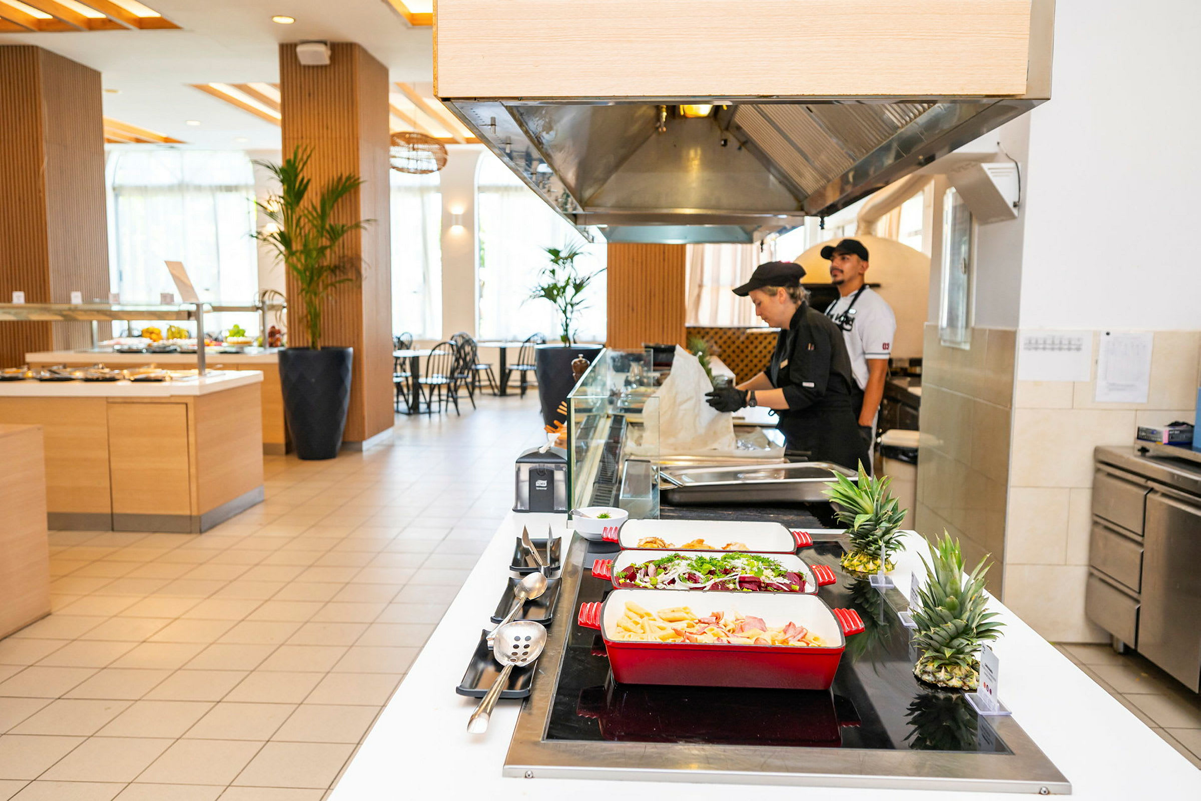 Bufféområde med mat upplagd i färgglada serveringsfat och kökspersonal vid arbetsstation. (AI-genererad text)