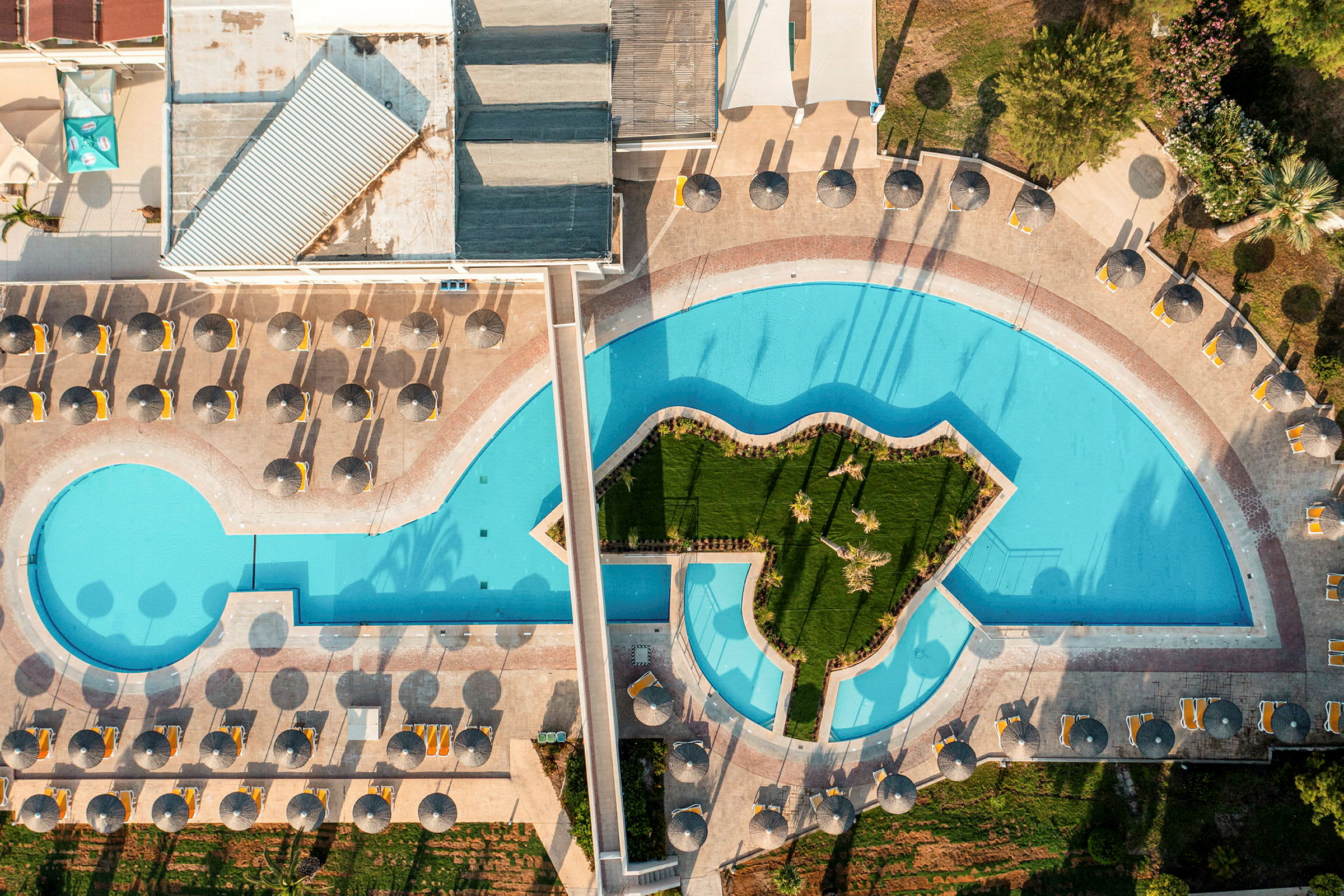 Flygfoto över en swimmingpool med en gräsbevuxen ö i mitten, solsängar och parasoller runtom. (AI-genererad text)