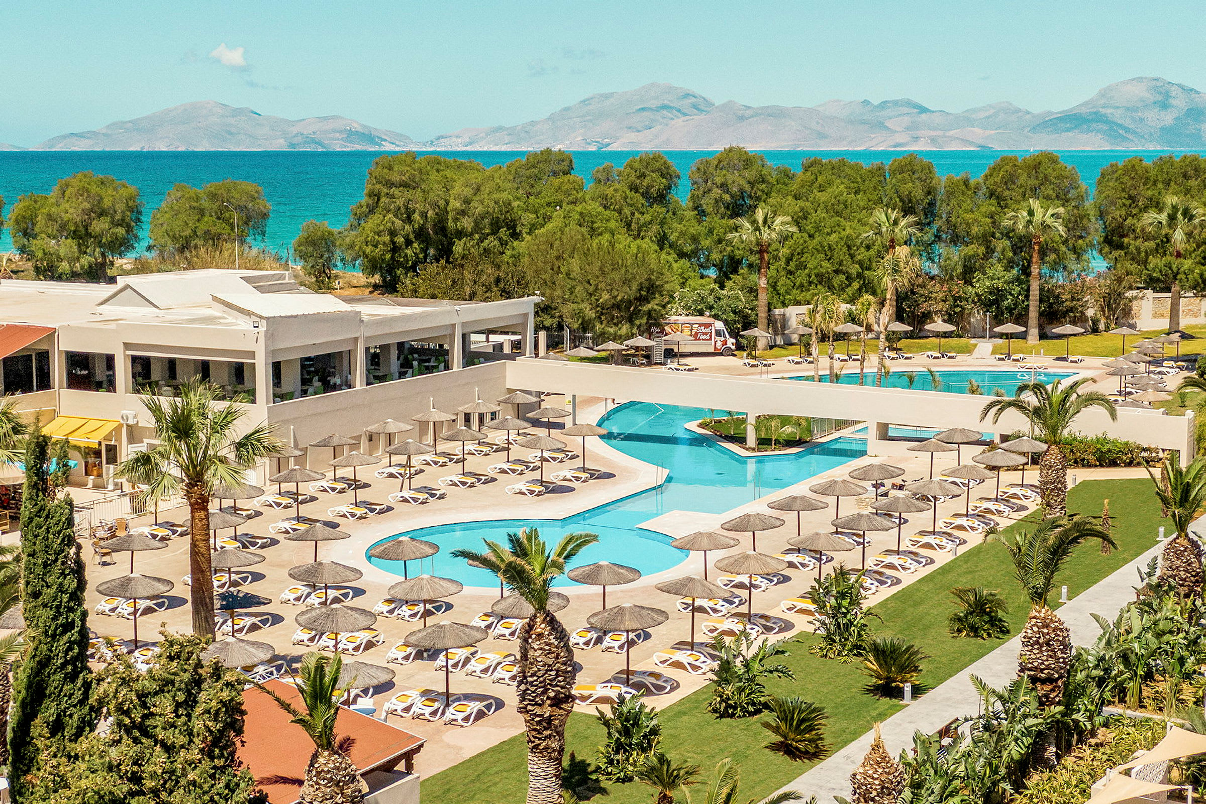 Hotellområde med pool, solstolar och parasoller, omgiven av grönska och med havet i bakgrunden. (AI-genererad text)