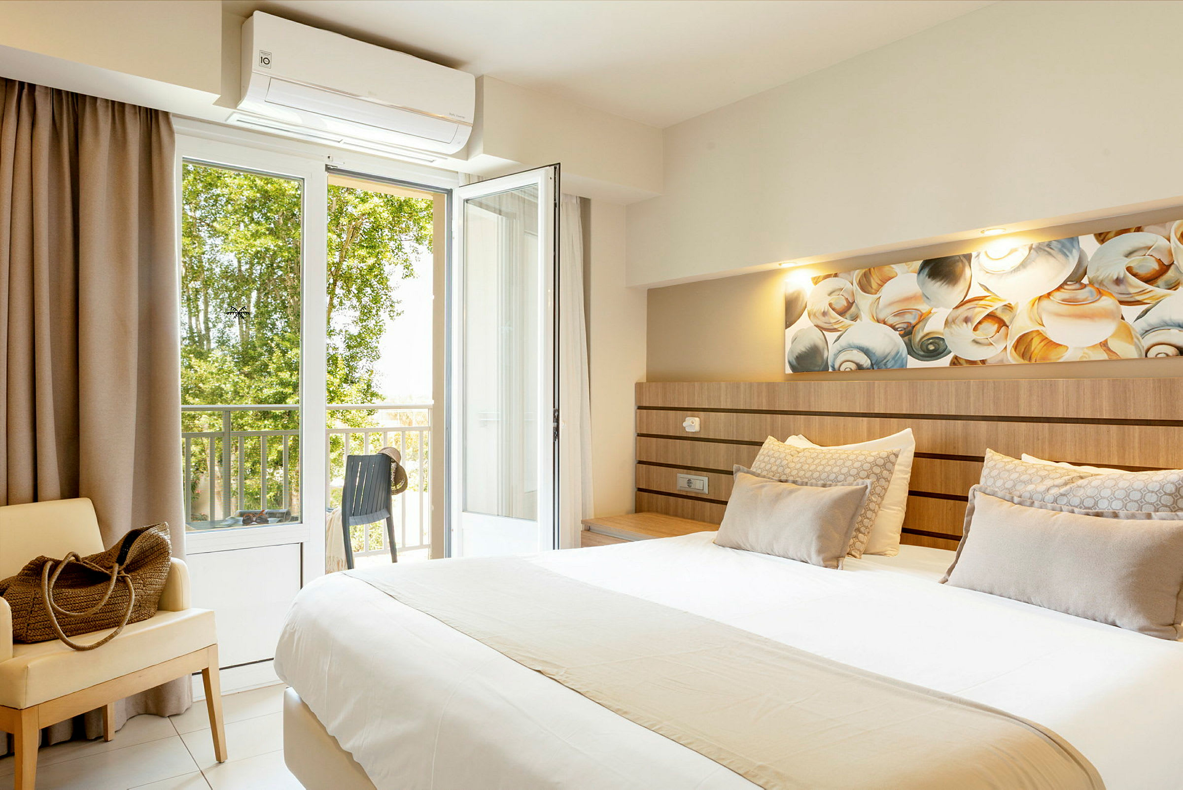 Ett hotellrum med en stor säng, sittmöbel, och öppen balkongdörr med utsikt mot grönska. (AI-genererad text)