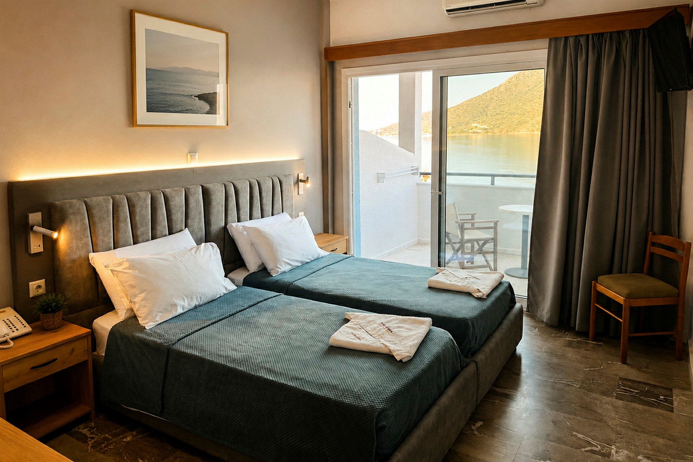 Hotellrum med två enkelsängar, sängbord och tavla på väggen. Balkong med utsikt över vatten och berg. (AI-genererad text)