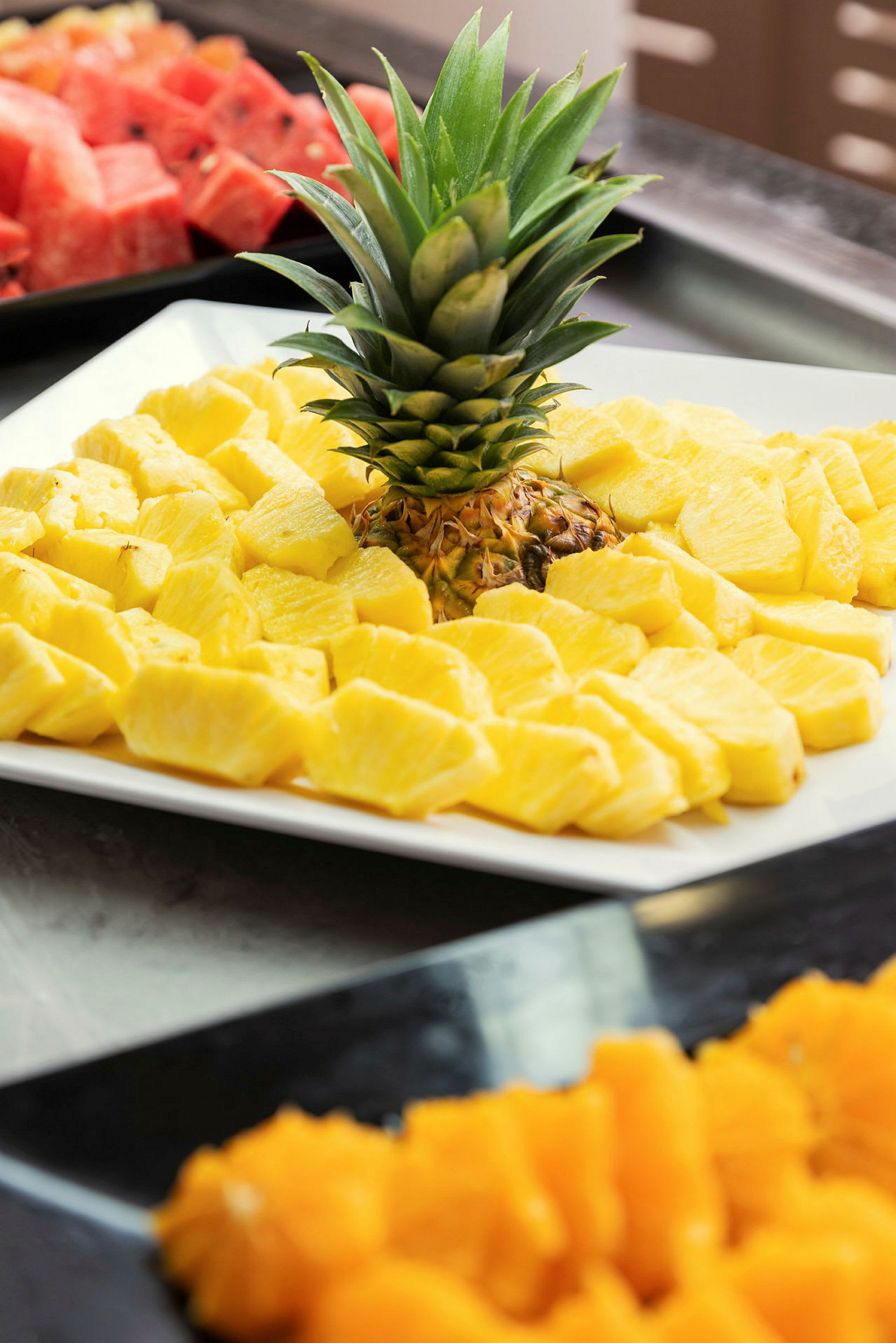 En närbild på en tallrik med skivade färska ananas och dess gröna krona, med andra frukter i bakgrunden. (AI-genererad text)
