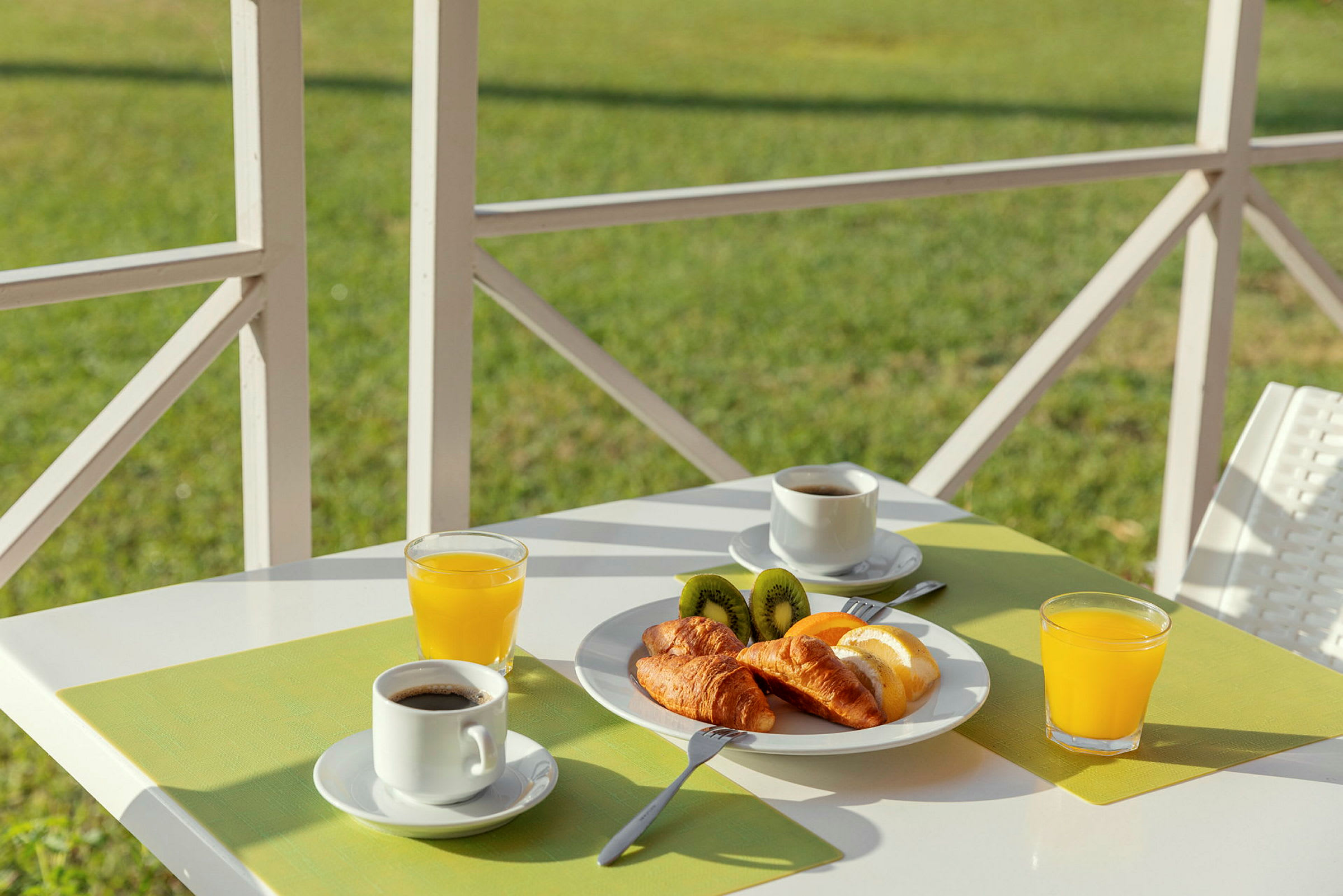 Frukost med kaffe, juice, croissanter och kiwi på en vit bord med grön bordstablett. (AI-genererad text)