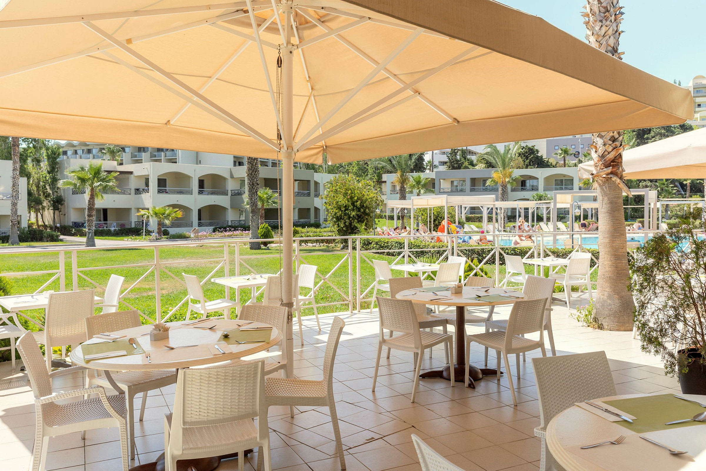 Utomhusrestaurang med möblerade bord nära en pool och omgivande byggnader. (AI-genererad text)