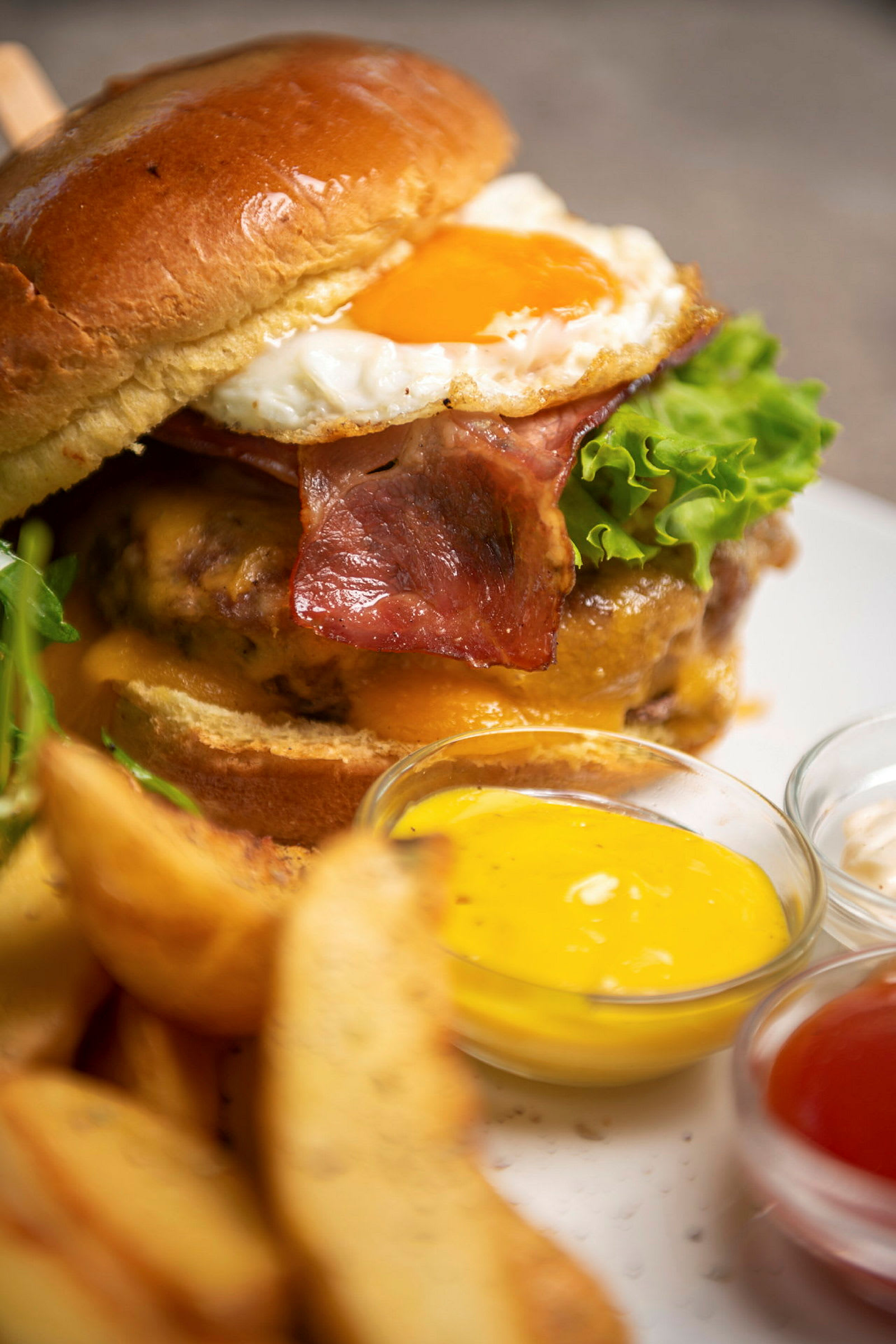 En hamburgare med bacon, ost, sallad och stekt ägg serveras med potatisklyftor och olika såser. (AI-genererad text)