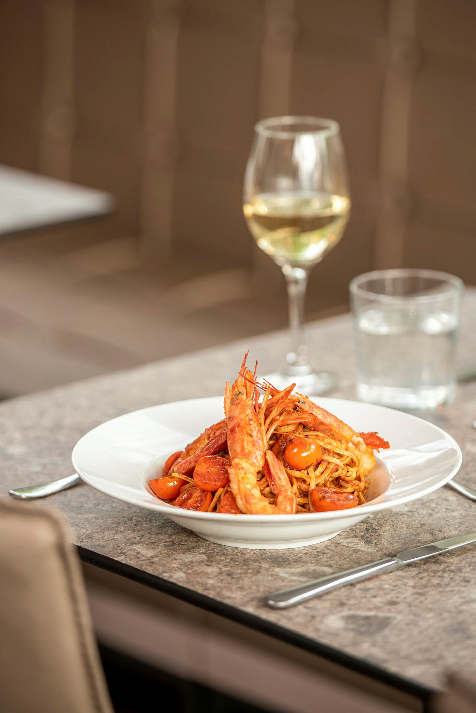 En tallrik med pasta, räkor och tomater står på ett bord. I bakgrunden syns ett glas vitt vin. (AI-genererad text)