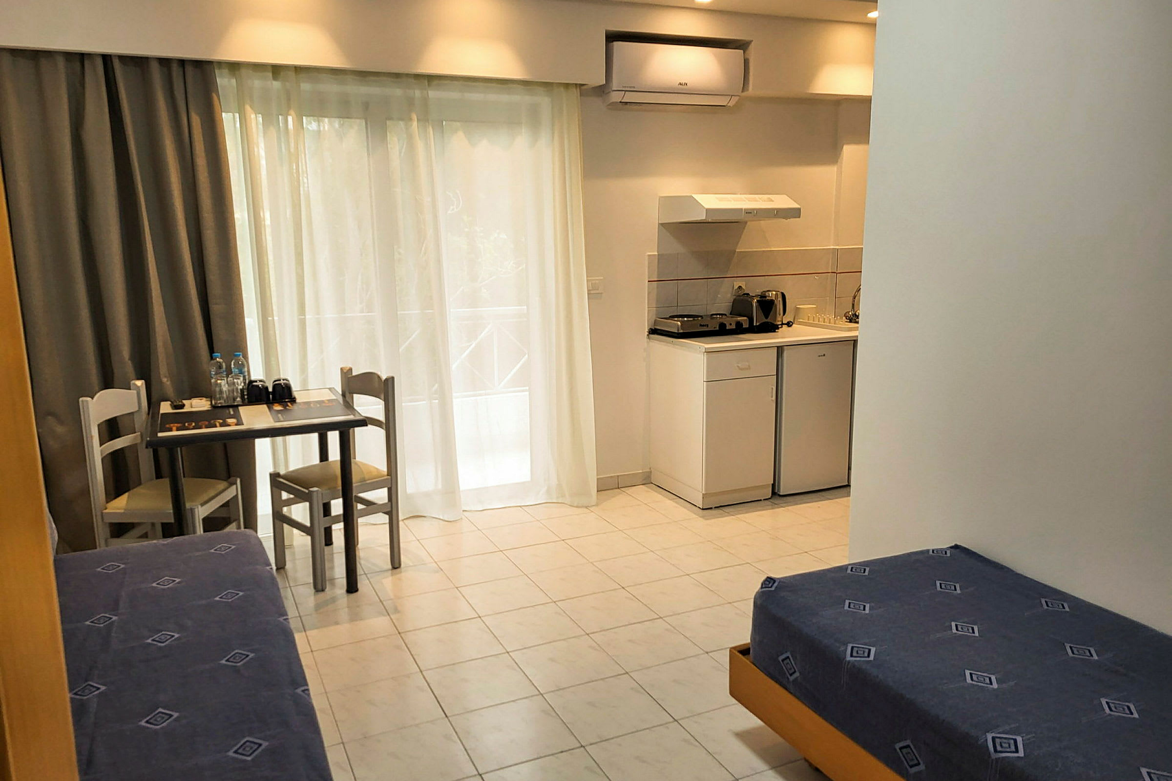 Ett hotellrum med enkla möbler inklusive sängar, ett litet bord med två stolar, samt ett pentry med kylskåp, spis och köksutrustning. (AI-genererad text)