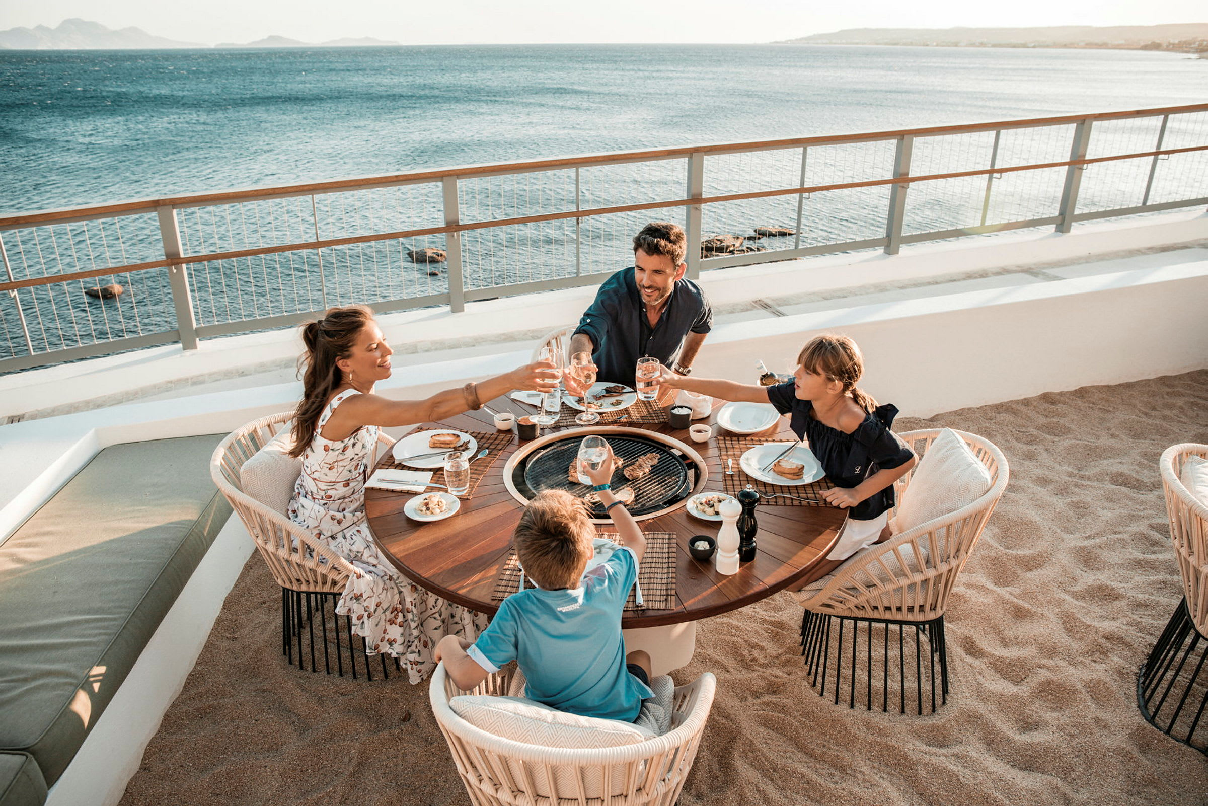 En familj sitter vid ett runt bord utomhus vid stranden och njuter av en måltid med utsikt över havet. (AI-genererad text)
