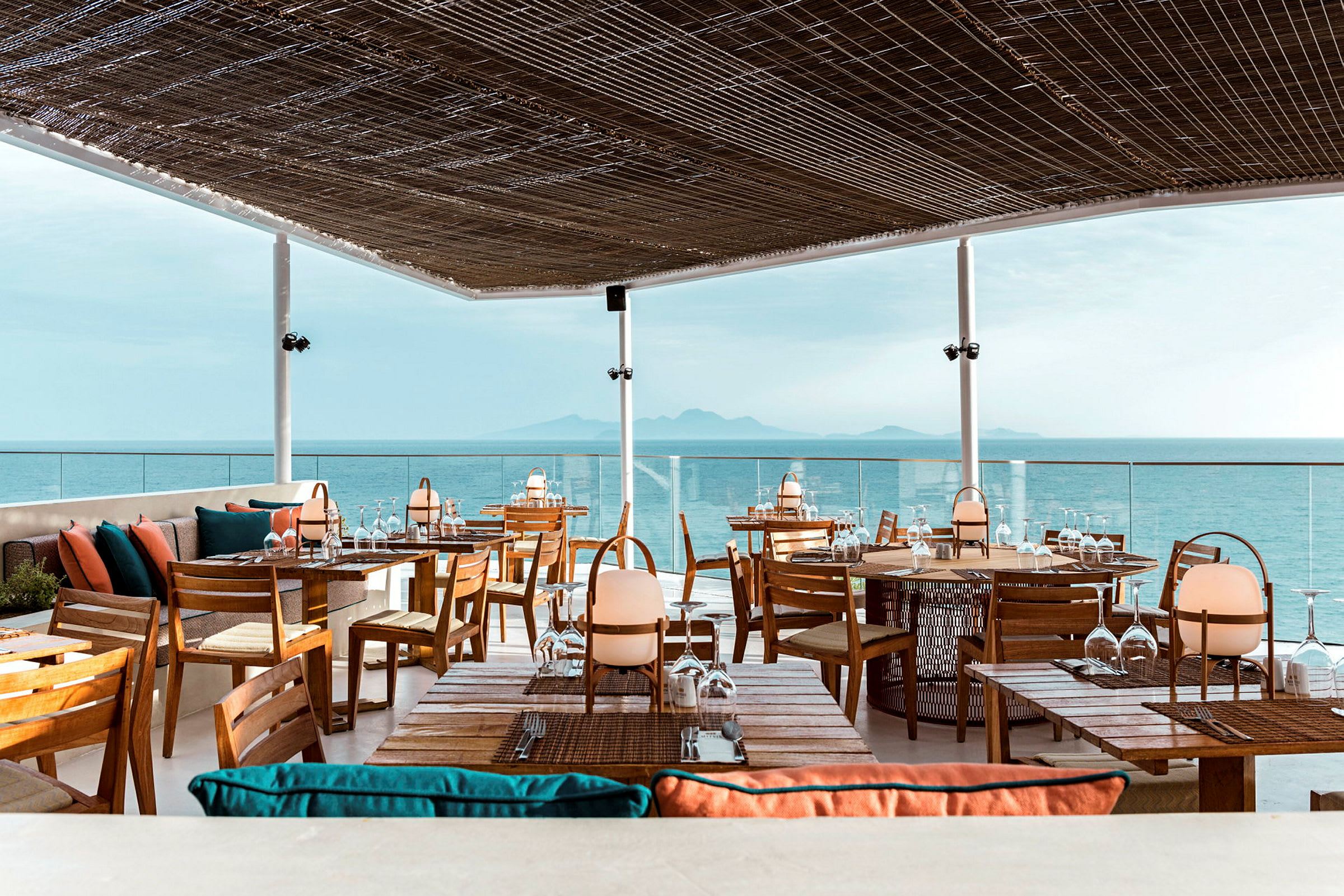 En restaurang med takterrass med utsikt över havet. Möblerna är av trä med färgglada kuddar, och taket består av ett flätat material som släpper in ljus. (AI-genererad text)