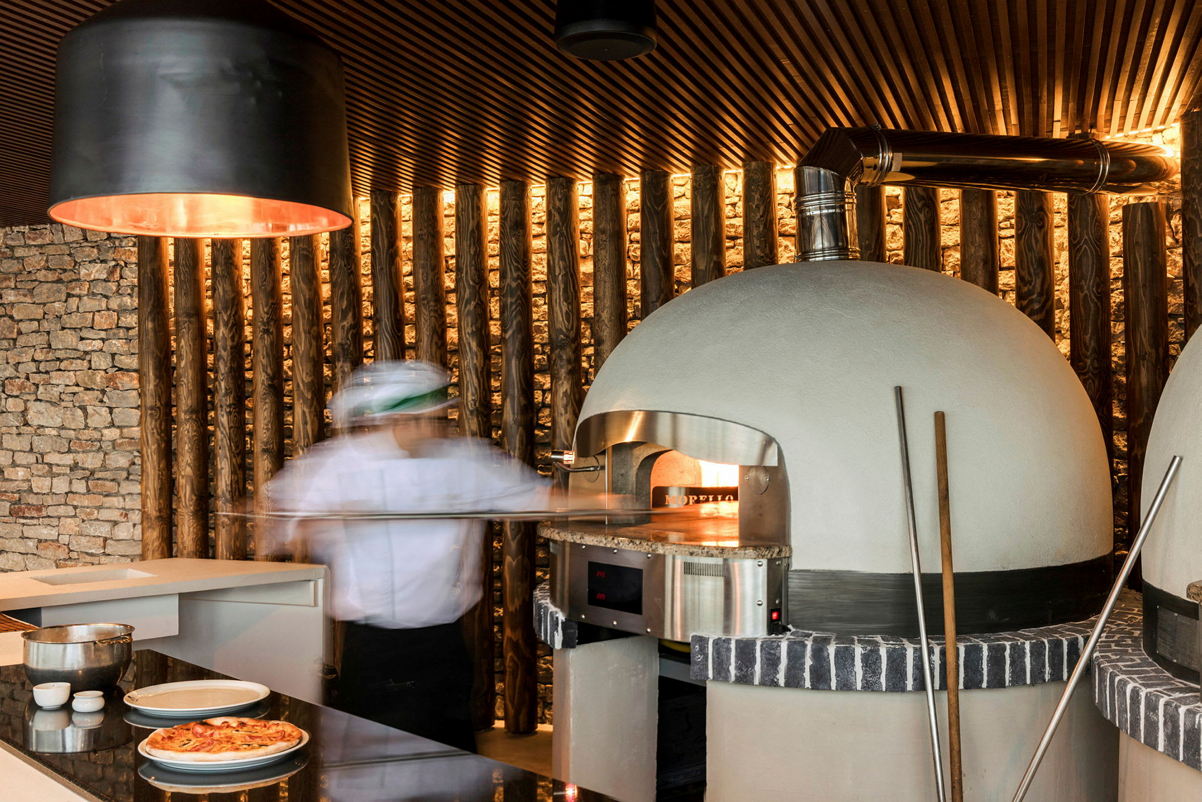 En pizzabagare framför en vedeldad ugn med ett modernt kök i bakgrunden. (AI-genererad text)