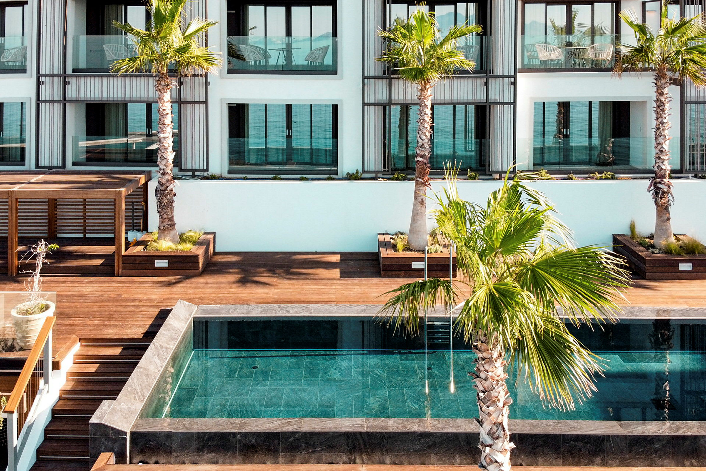 En pool omgiven av träplattformar och palmer framför ett modernt hotell. (AI-genererad text)