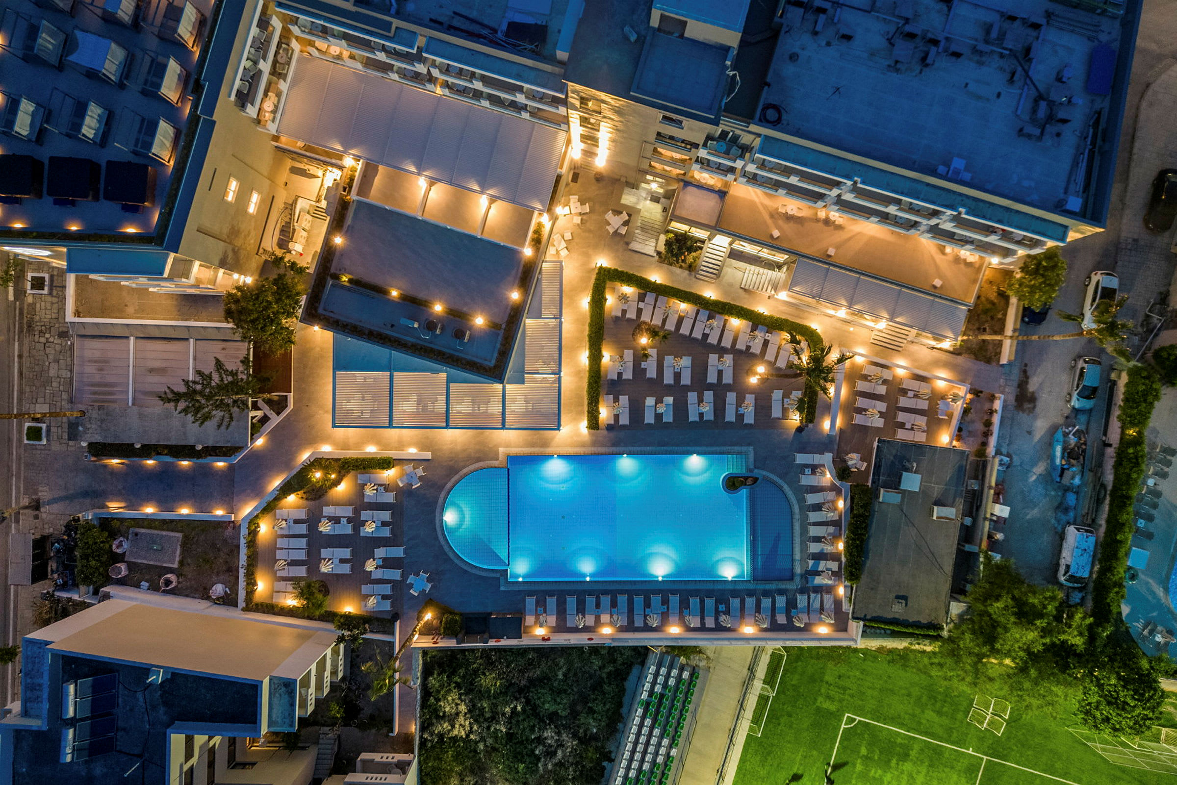 Flygfoto av hotellområde med pool, solstolar och grönområden, belyst på kvällen. (AI-genererad text)