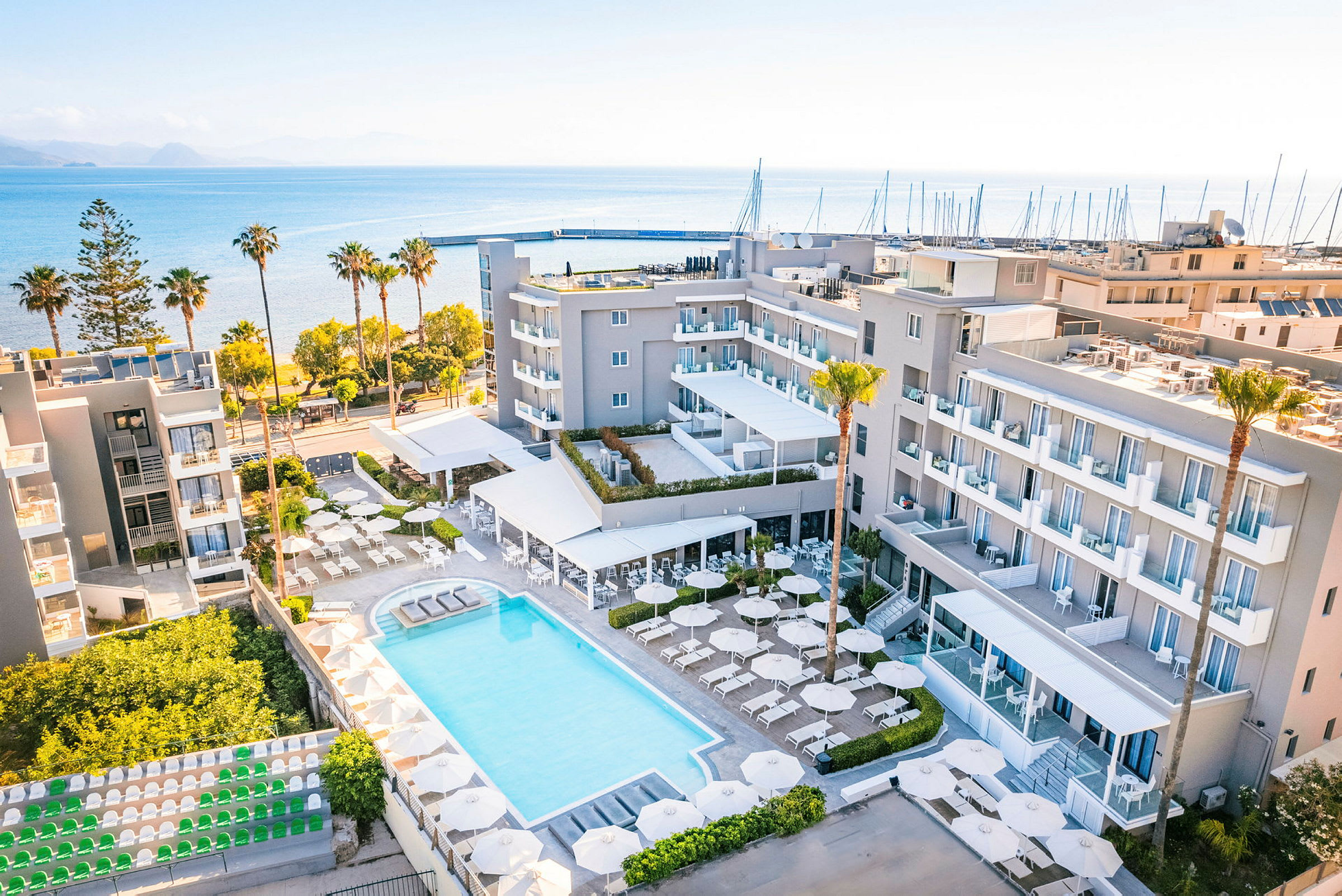 Ett hotellområde med pool, parasoller och solstolar omgivna av byggnader. Hotellet ligger nära havet med en strand och en småbåtshamn i bakgrunden. (AI-genererad text)