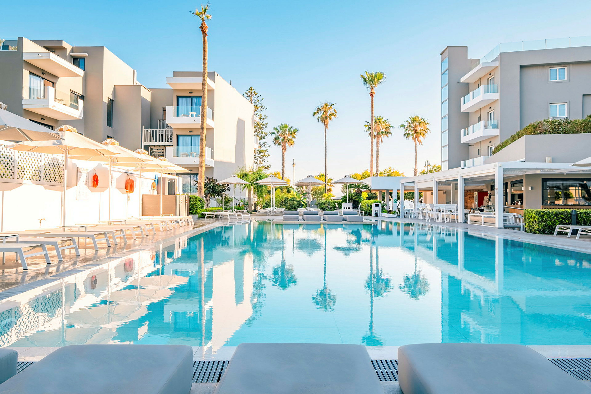 En modern hotellbyggnad med en upplyst pool framför, flankerad av solstolar och parasoll. (AI-genererad text)