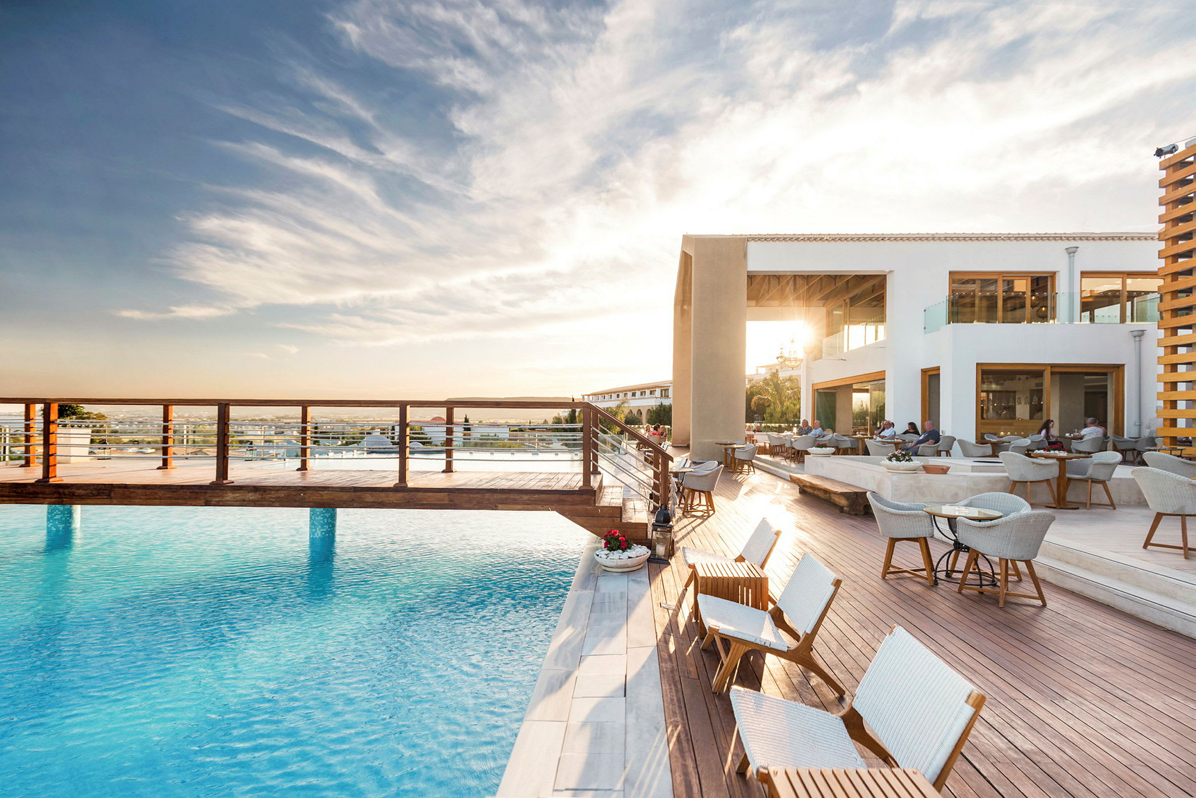 En utomhus pool med trädäcket och sittmöbler, ett modern byggnad med kvällssol i bakgrunden. (AI-genererad text)