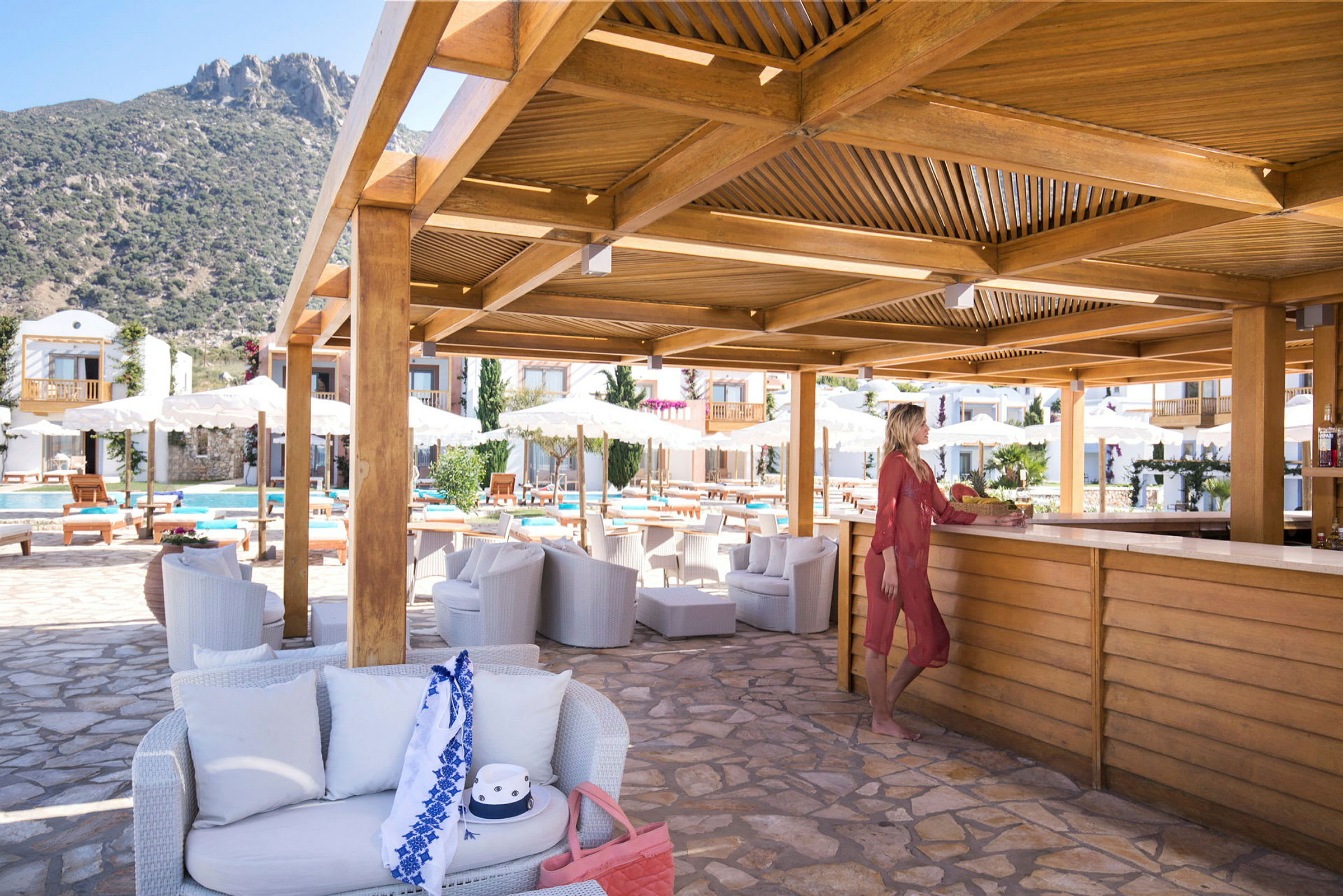 Poolområde med bar och sittplatser under ett träpergola. Berg i bakgrunden. (AI-genererad text)