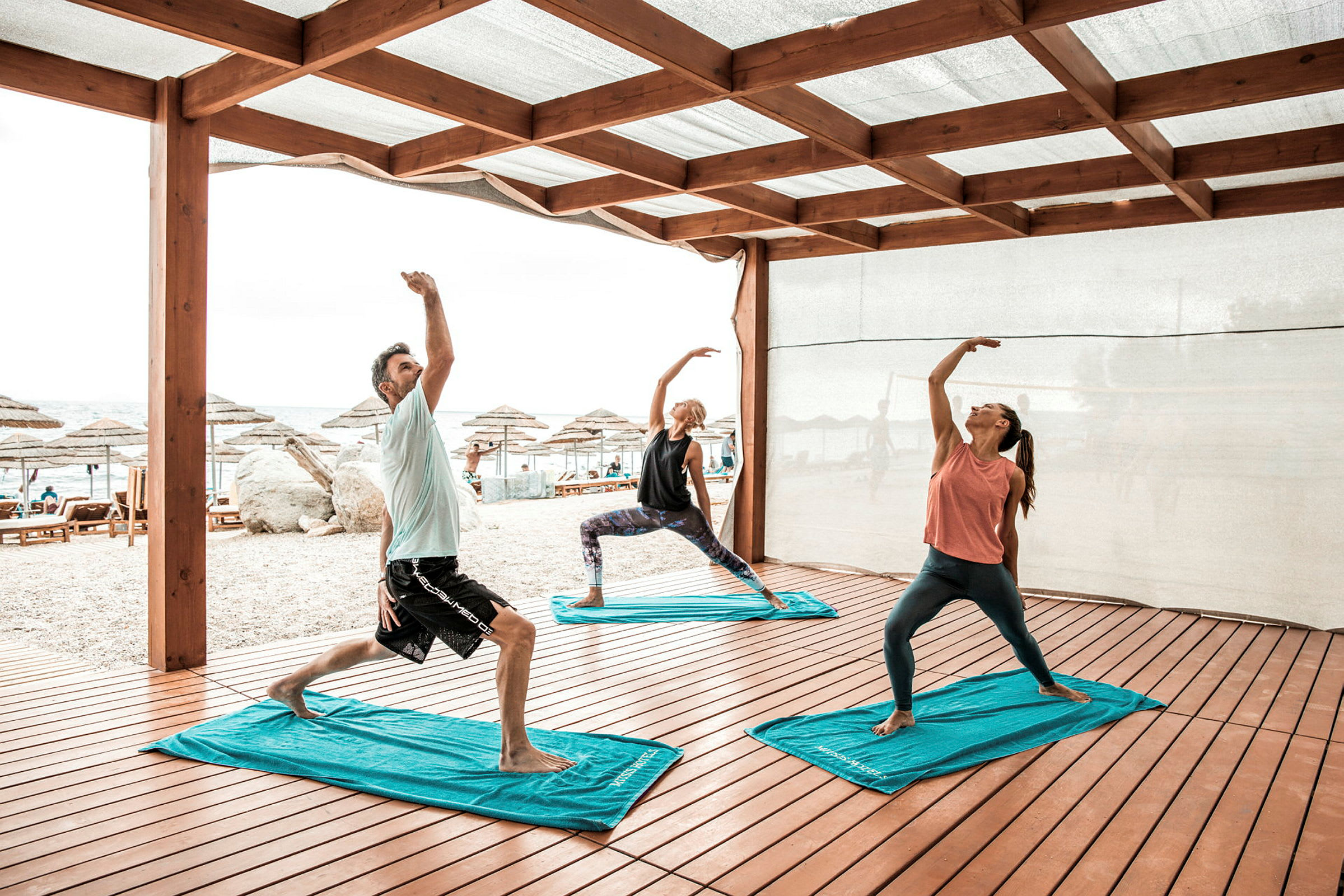 Tre personer utför yogaövningar under ett träskydd nära en strand. (AI-genererad text)